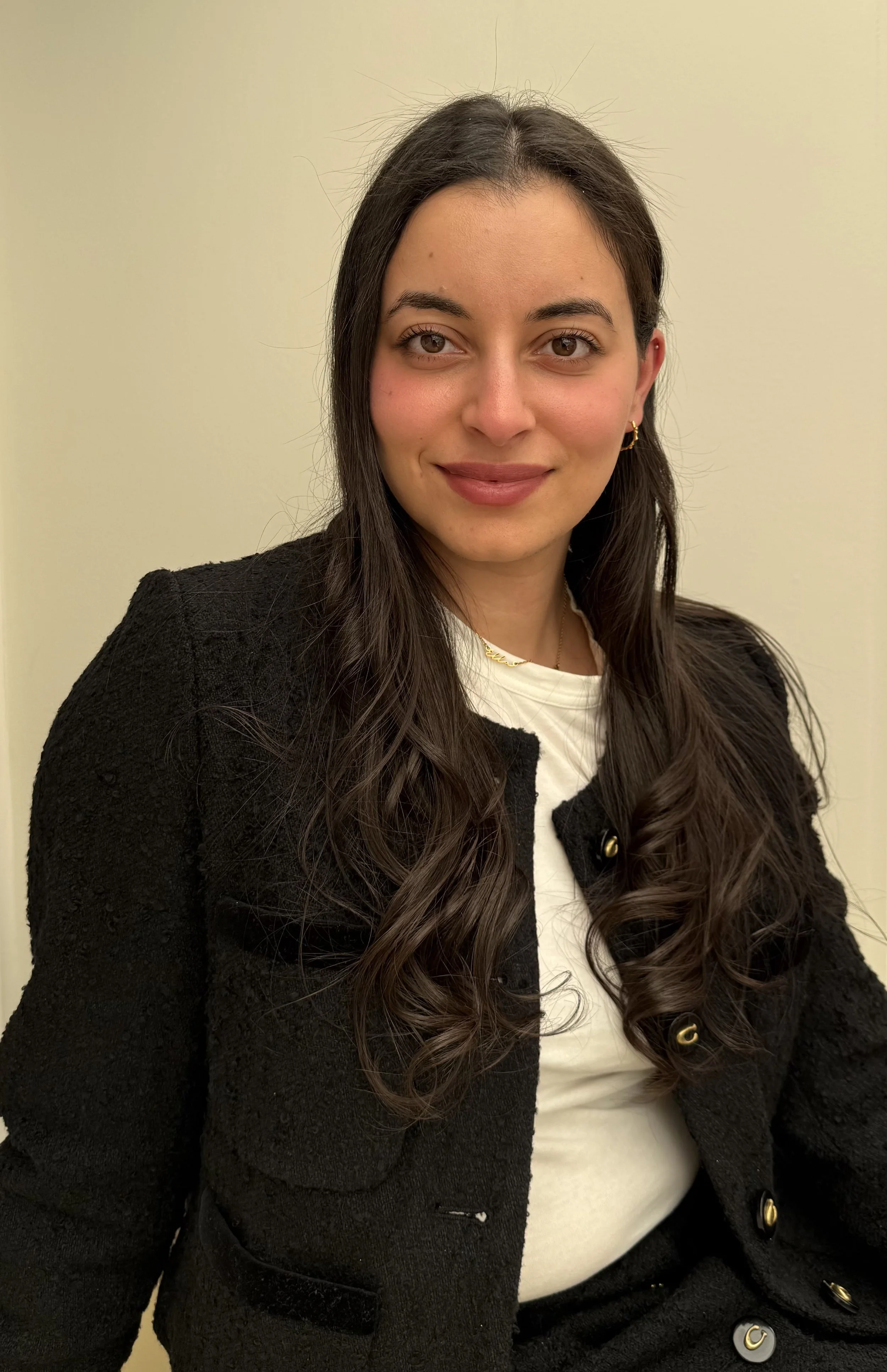 A woman with long dark hair, wearing a black textured blazer over a white top, smiling at the camera, against a plain light-colored background.