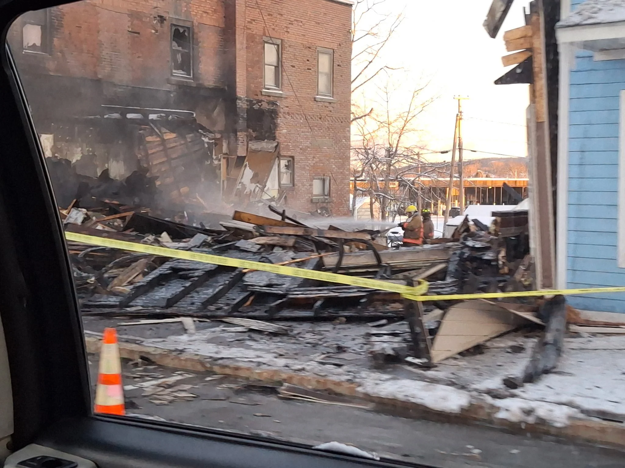 Debris from a building fire with firefighters in the background, yellow caution tape, and orange safety cone in a snowy scene.