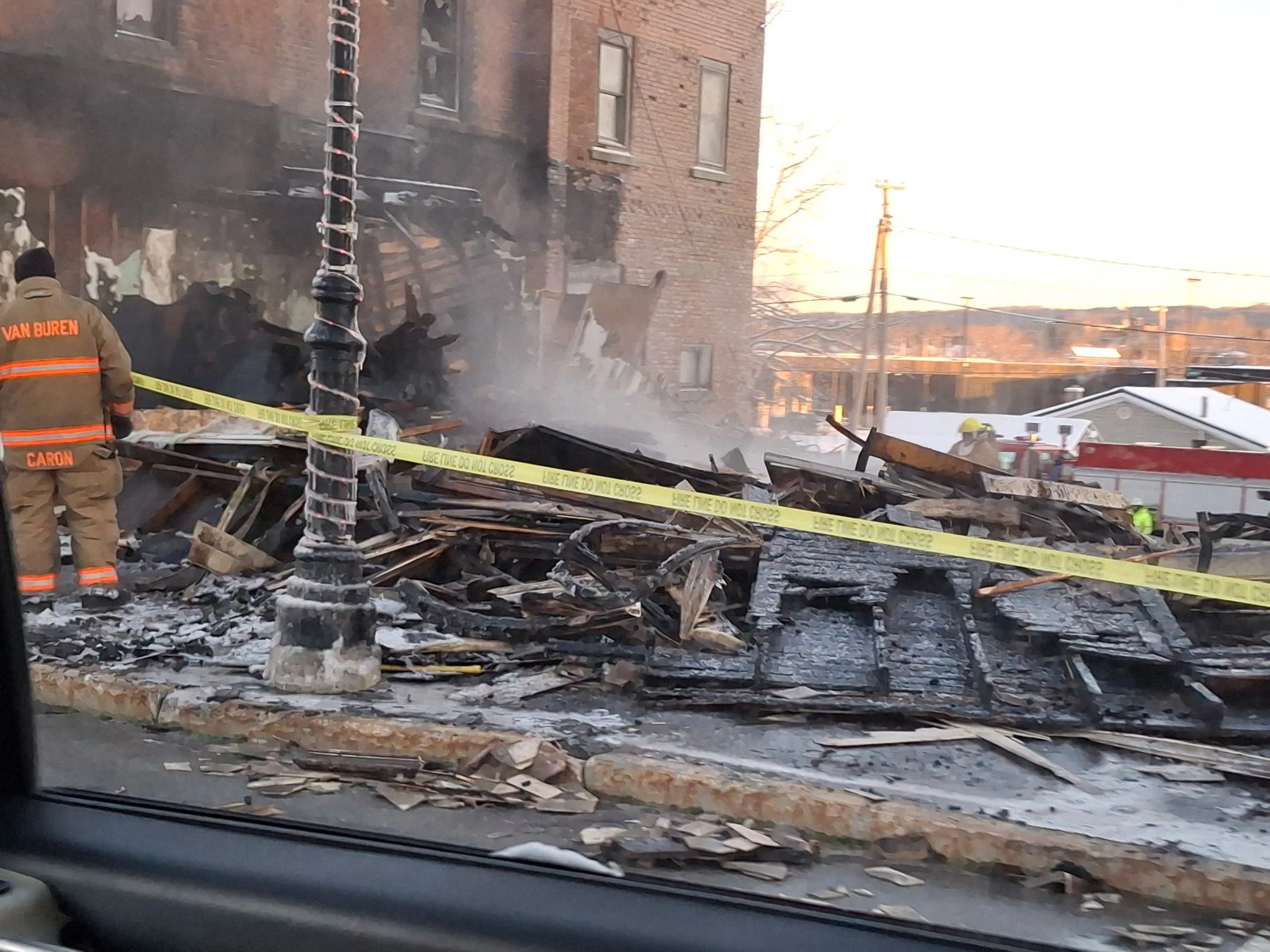 Firefighters investigate the aftermath of a building fire with damaged and charred debris, yellow caution tape, and smoke, in a residential area near a brick building.