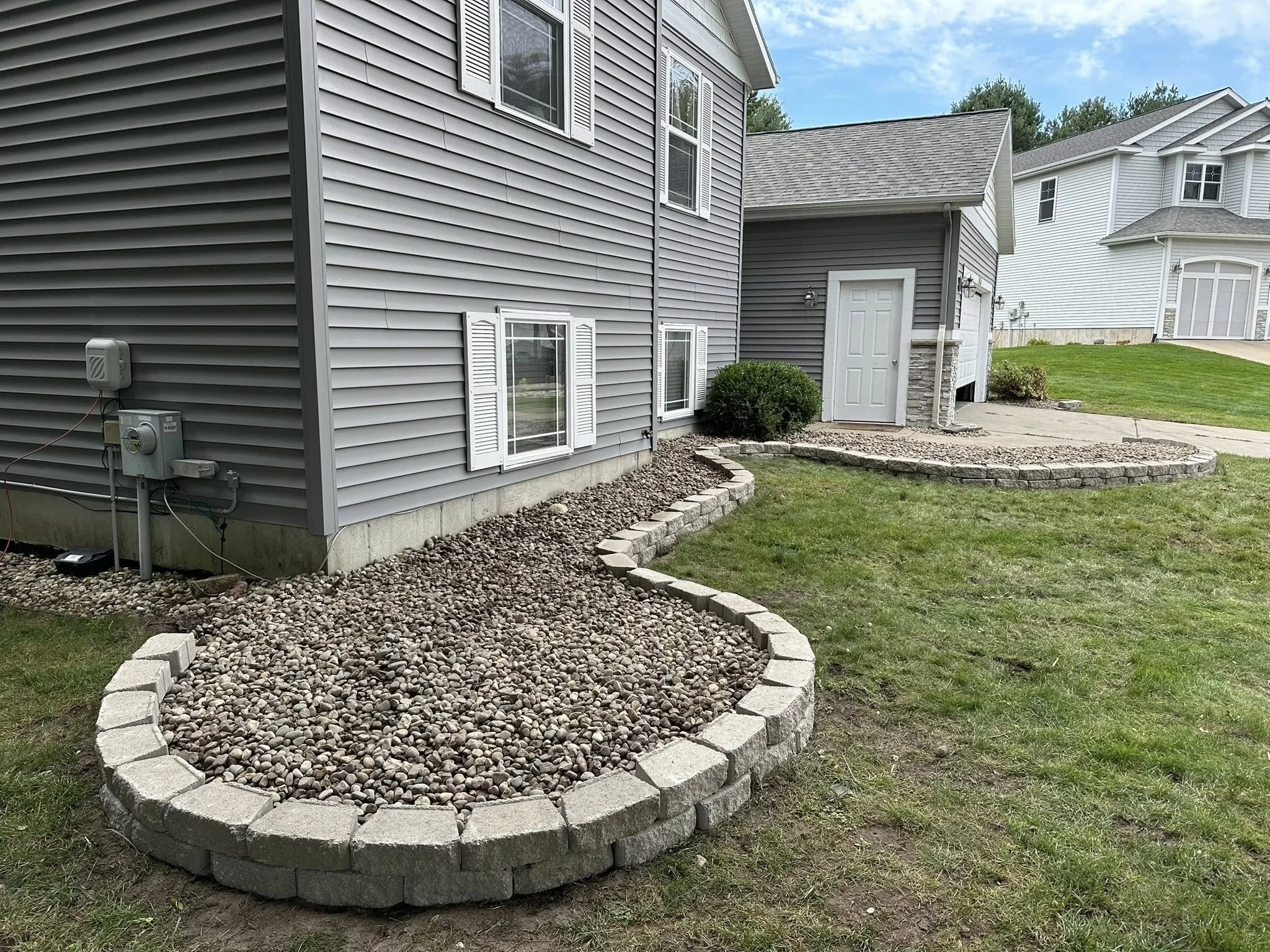 Backyard of a house with a gravel garden bed bordered with stone blocks, next to a gray house with white shutters and a smaller building with a door, and other homes in the background.