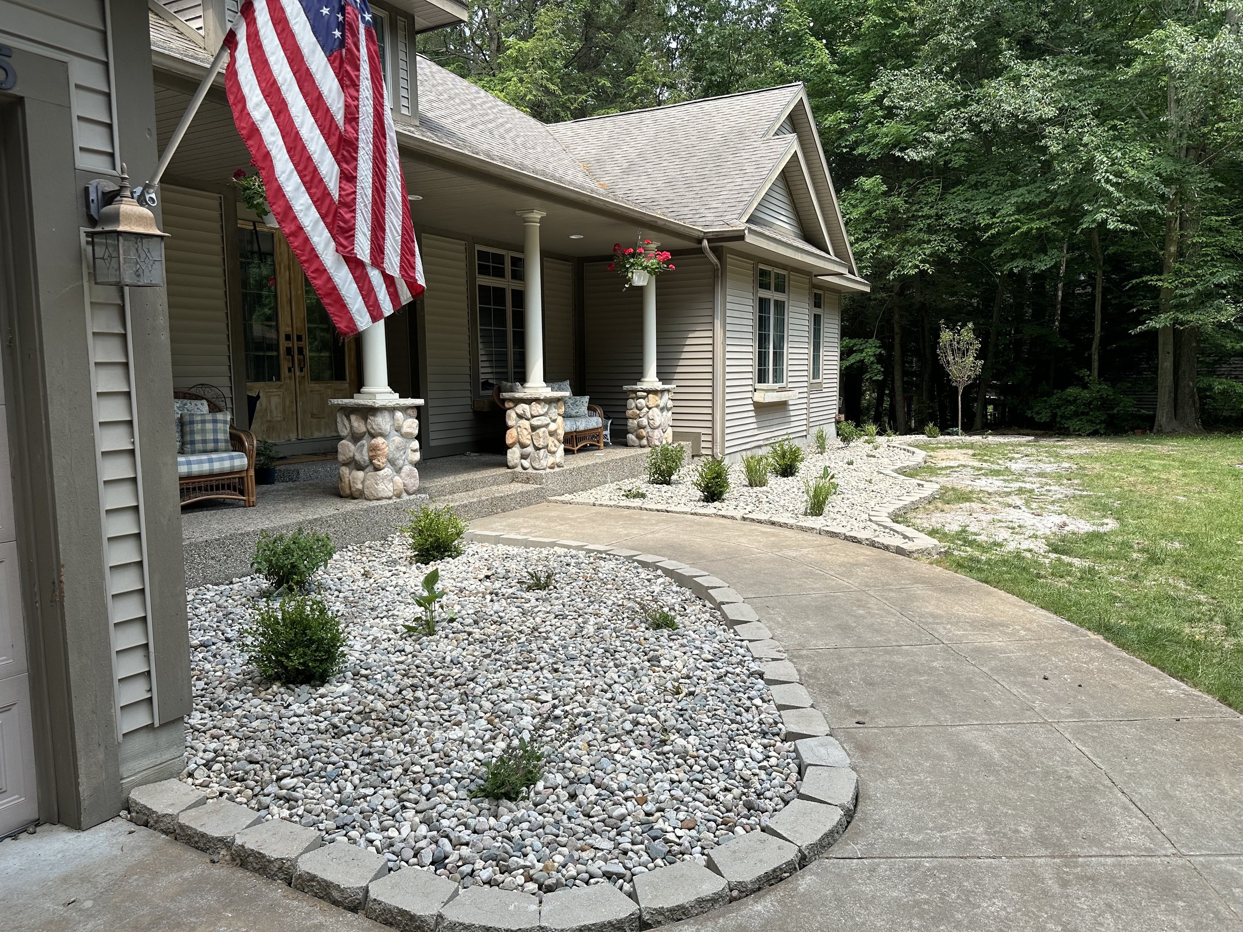 Brand new landscaping by Royle Exterior. Front porch of a house with American flag, rocking chairs, hanging flower pot, and a curved stone and concrete pathway with a garden bed filled with small plants and rocks.