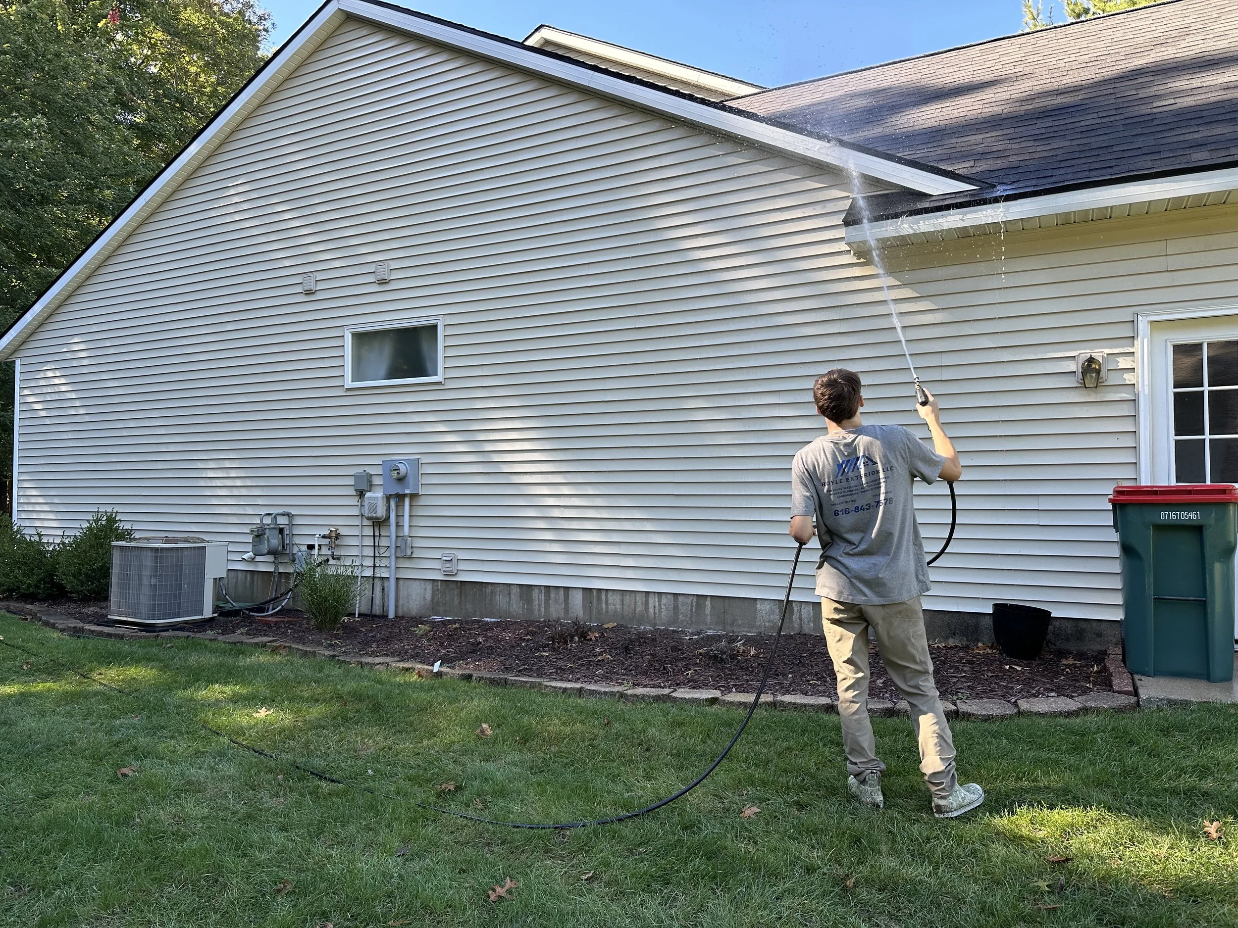 A man pressure washing the side of a house with white vinyl siding. There is a green trash bin nearby, and the house has a small window and some outdoor utility boxes and an air conditioning unit. The lawn is green and well-kept.