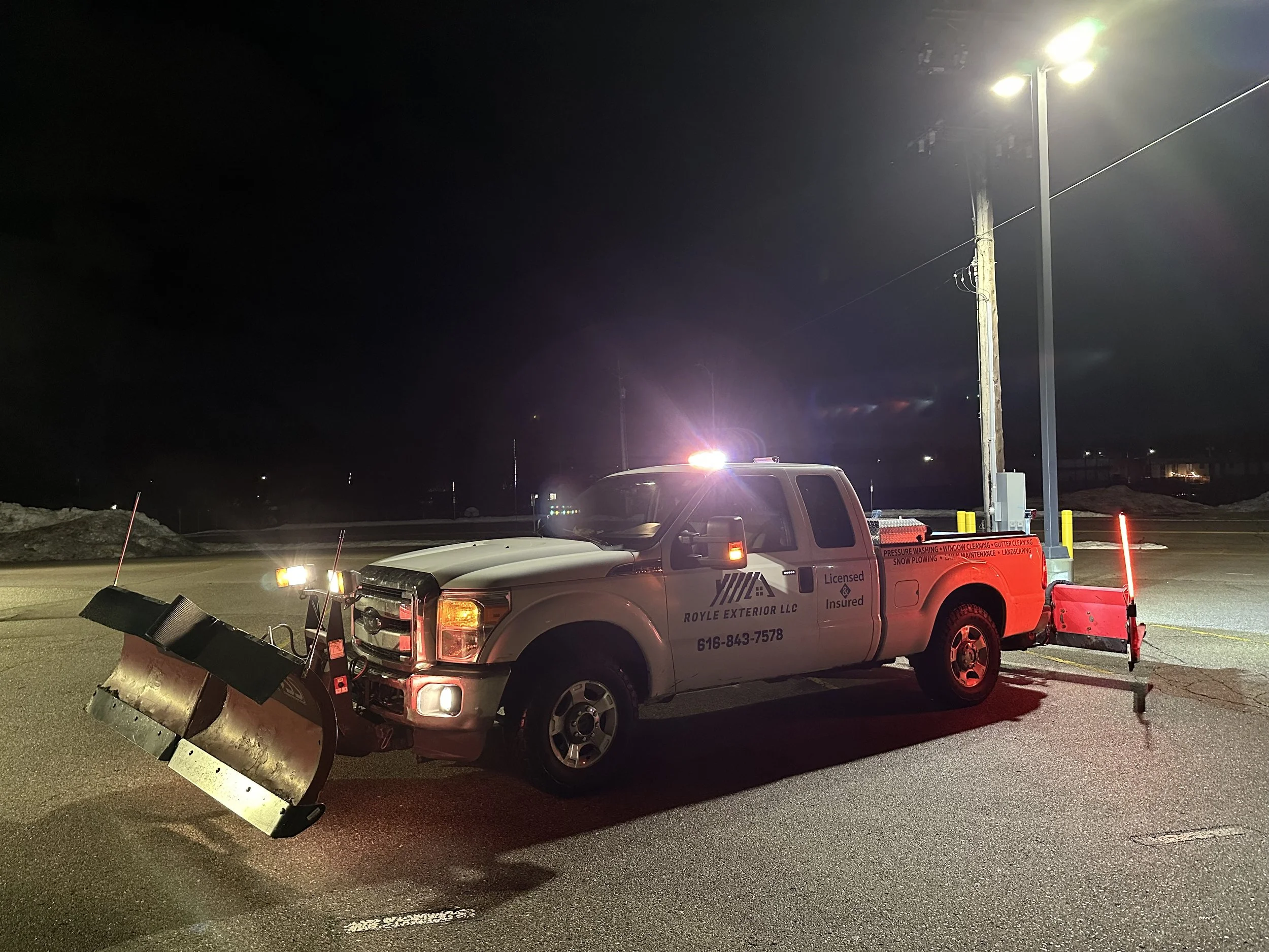 A utility truck marked 'Royle Exterior LLC' with snow removal equipment attached, parked on a road at night under streetlights, with the truck's roof lights on.