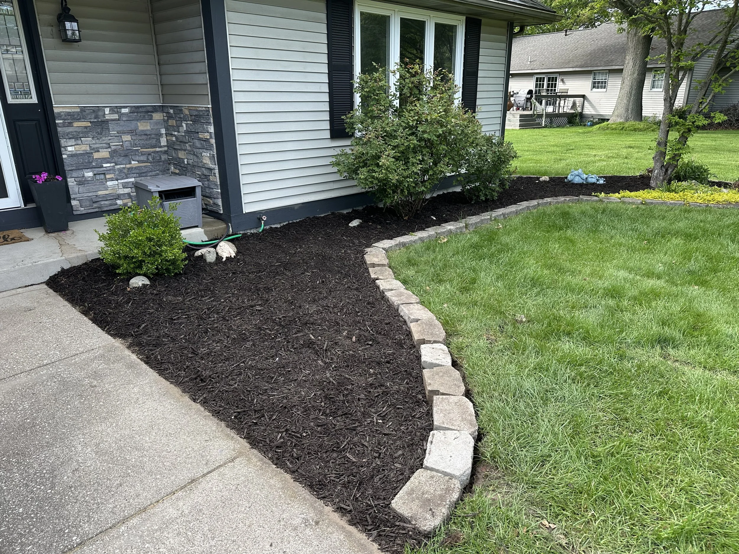 A landscaped front yard with a flower bed bordered by gray stone bricks, filled with dark mulch and small bushes. The house exterior features beige siding, a stone accent wall, black shutters, and a black lamp. There is a small gray storage box near the house and a garden hose connected to a spigot. The lawn is green and well-maintained, with a large tree in the background and neighboring houses visible.