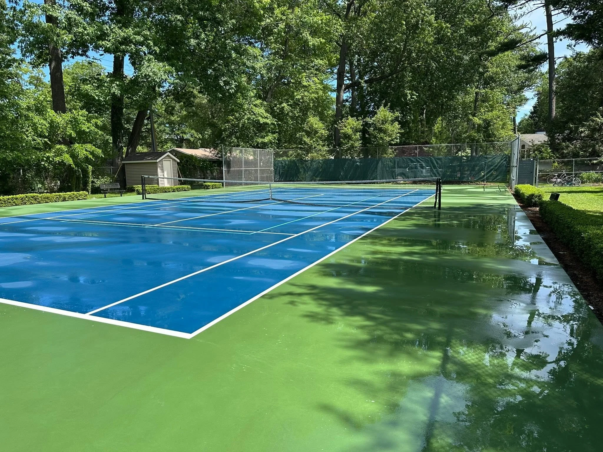 A blue tennis court surrounded by green trees and a chain-link fence with a small shed and bicycles in the background.