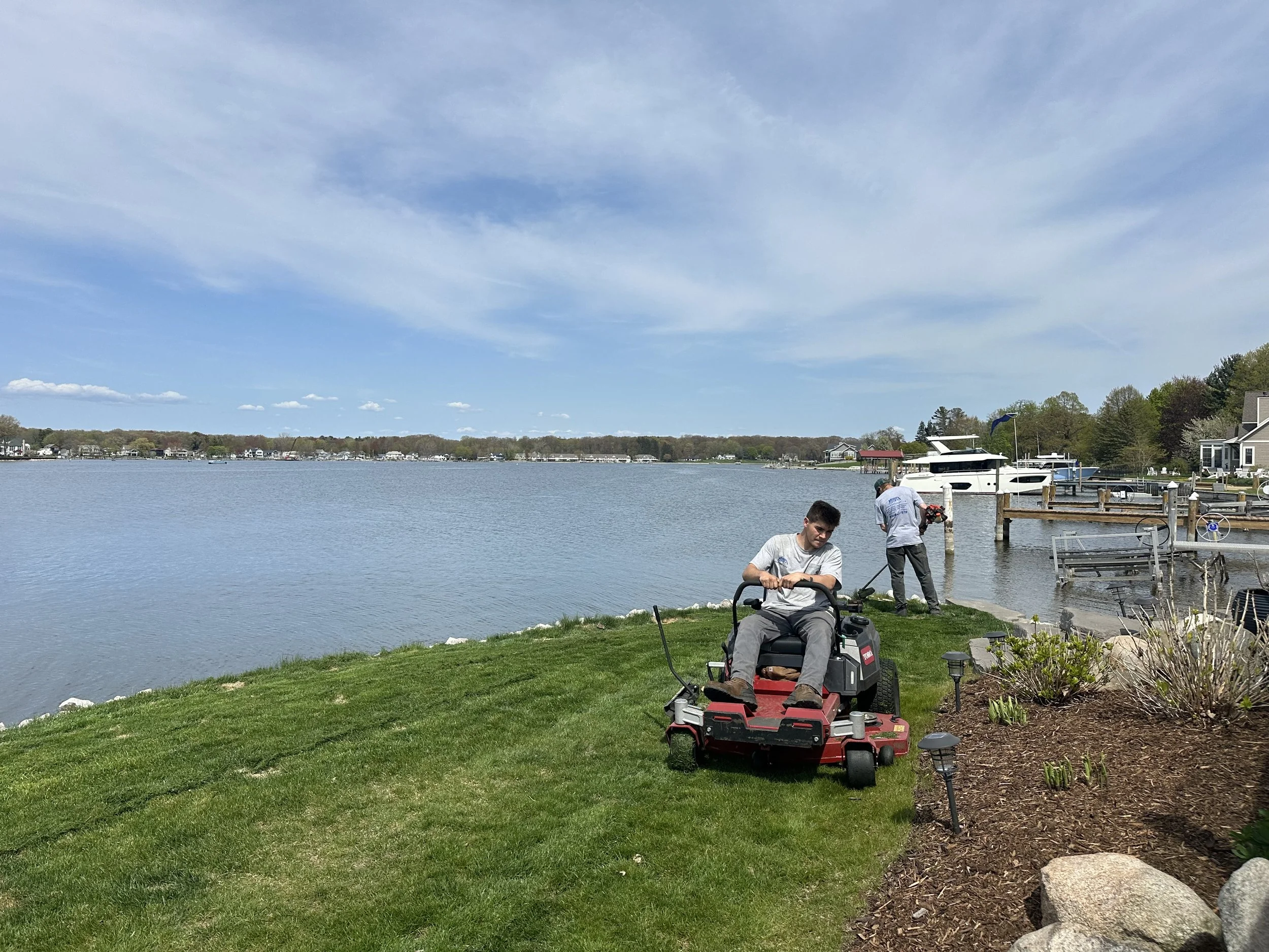 A young man mowing the lawn near a lake with two other men fishing on the dock in the background. There are houses along the shoreline, and the sky is partly cloudy.
