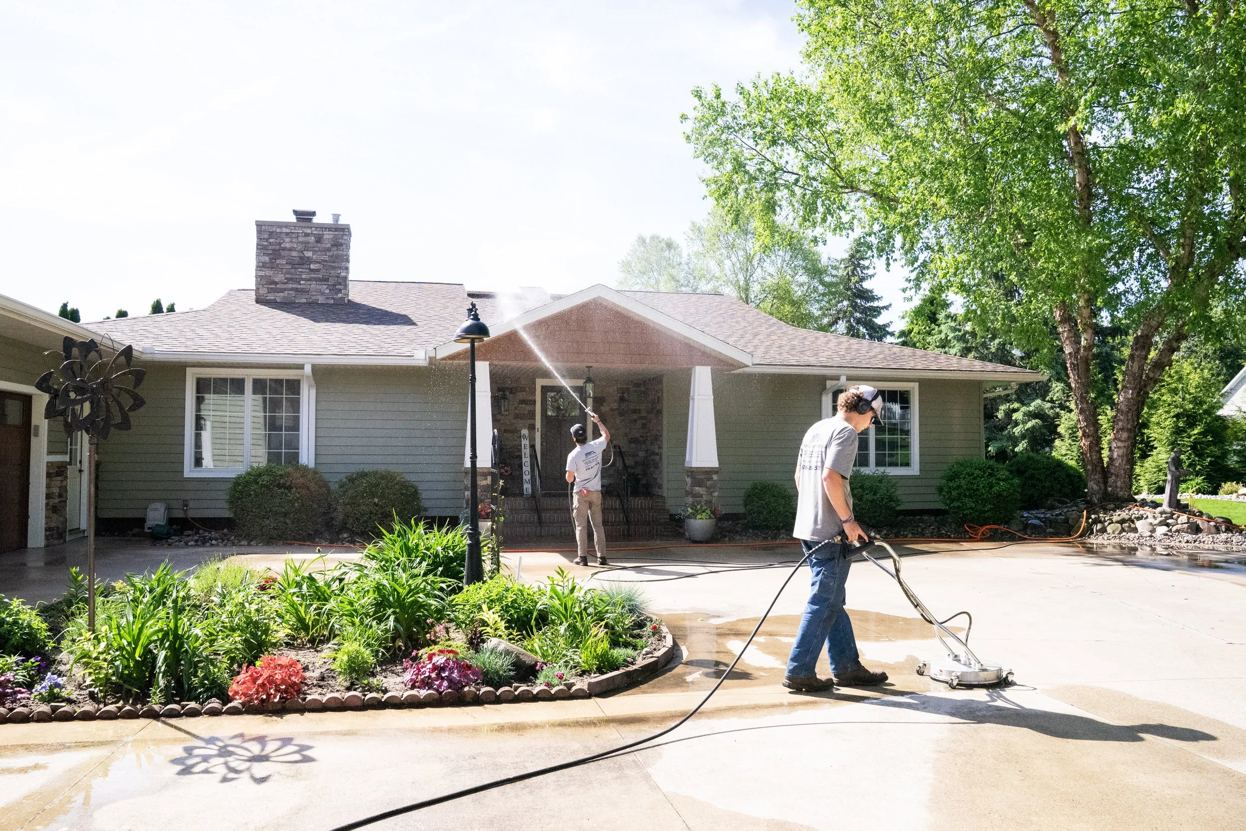 Royle Exterior team members pressure washing the driveway of a house with a garden and trees in the background on a sunny day.