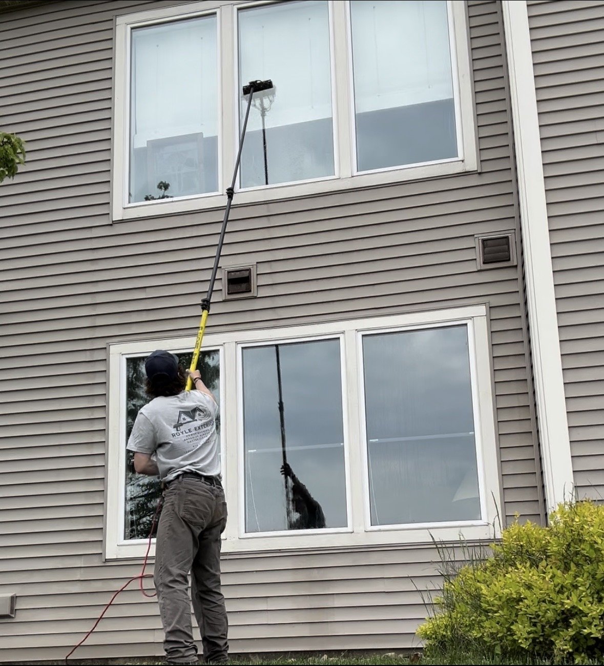 Royle Exterior team member cleaning window on a grey, multi-story building using extendable tool with a sponge or cloth at the end.
