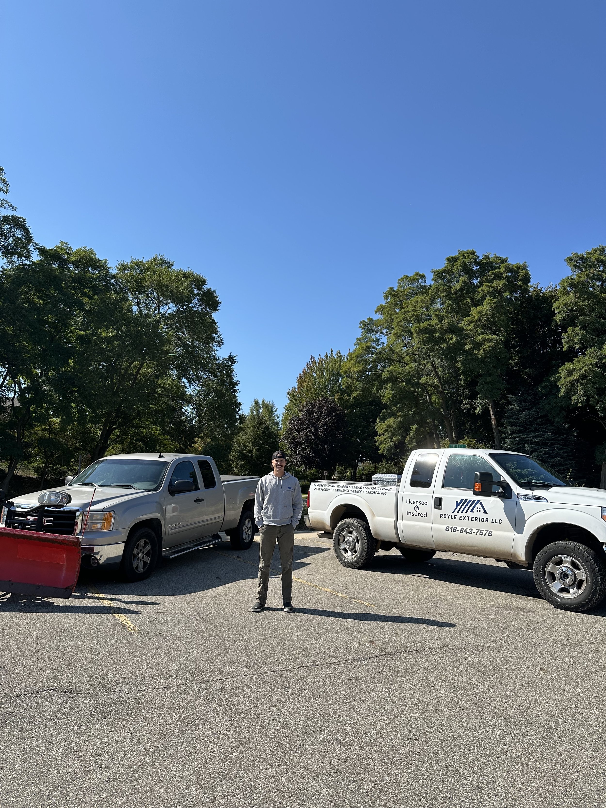 A man standing in a parking lot between two pickup trucks, one with a snow plow attached, surrounded by trees under a clear blue sky.