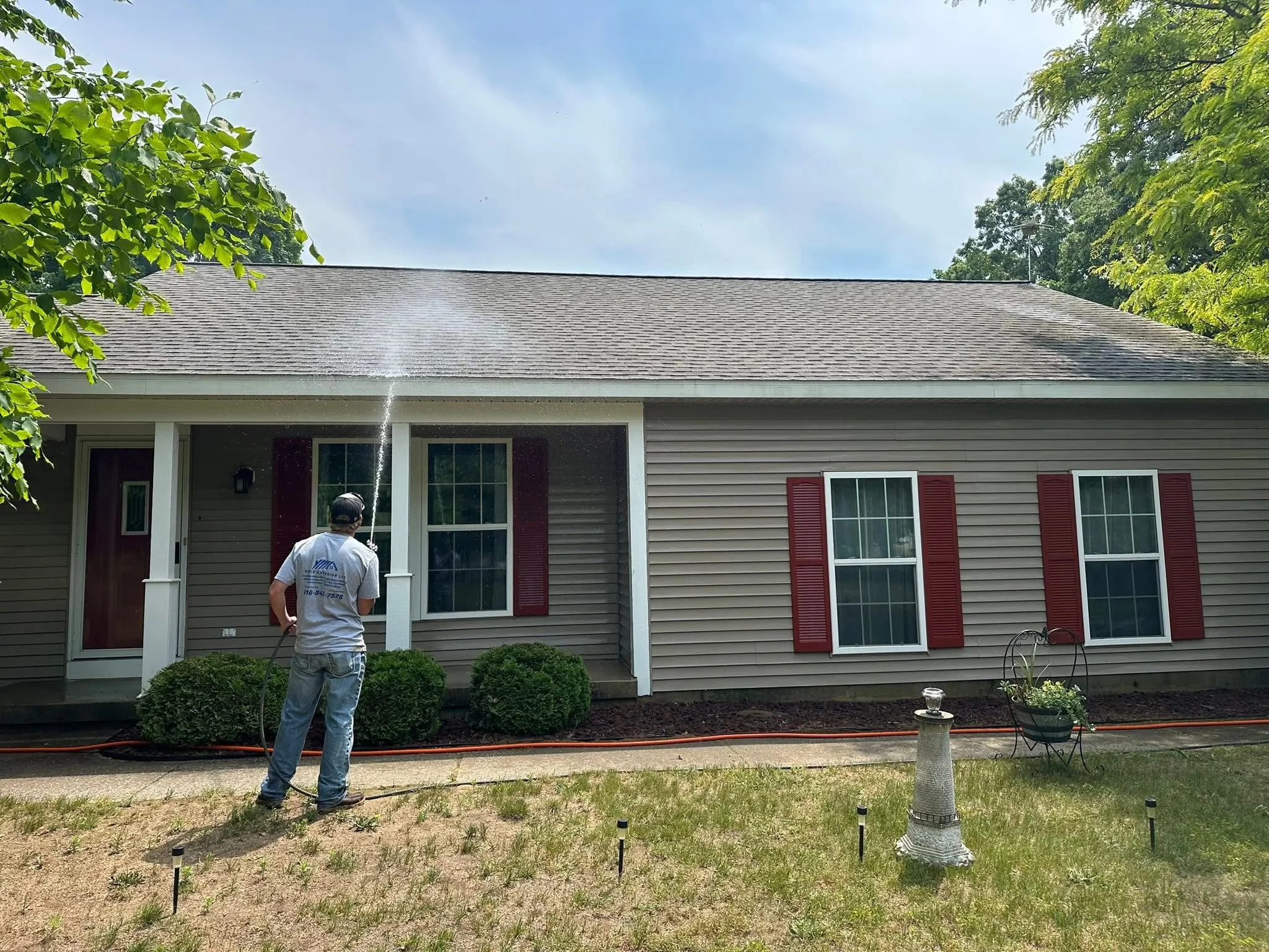 A person watering the front yard of a beige house with red shutters on a sunny day.
