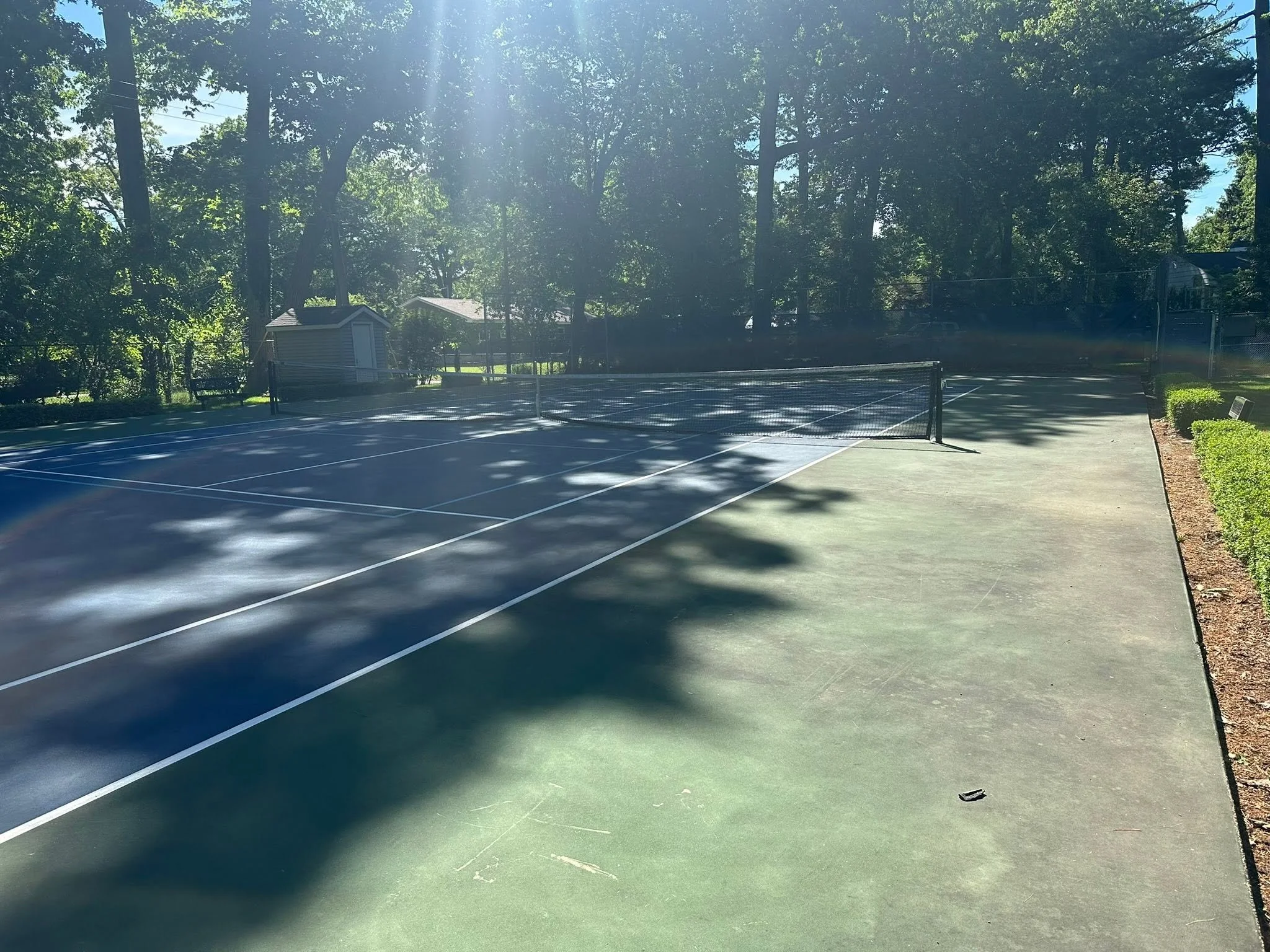 An outdoor tennis court surrounded by trees with sunlight streaming through the leaves.