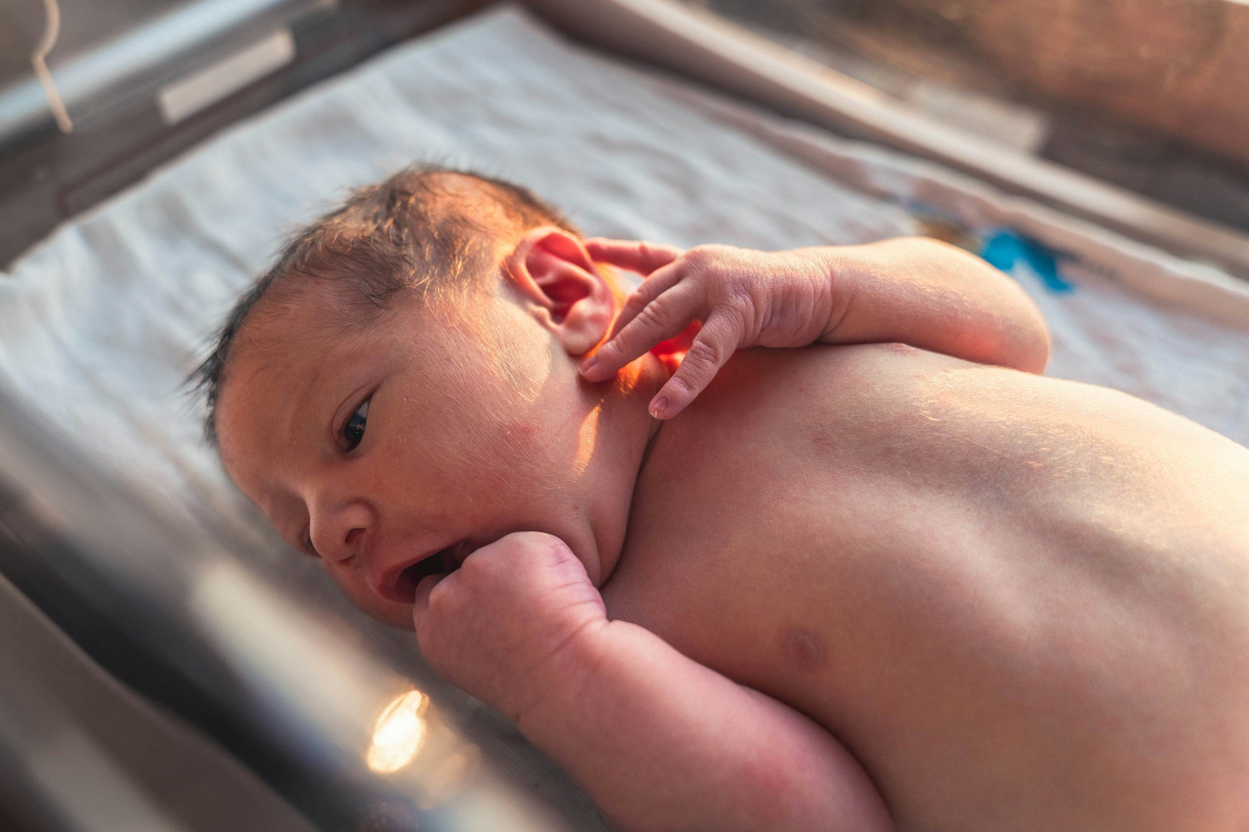 A newborn baby lying on a hospital bed with a hand touching its face, in the morning sunlight.
