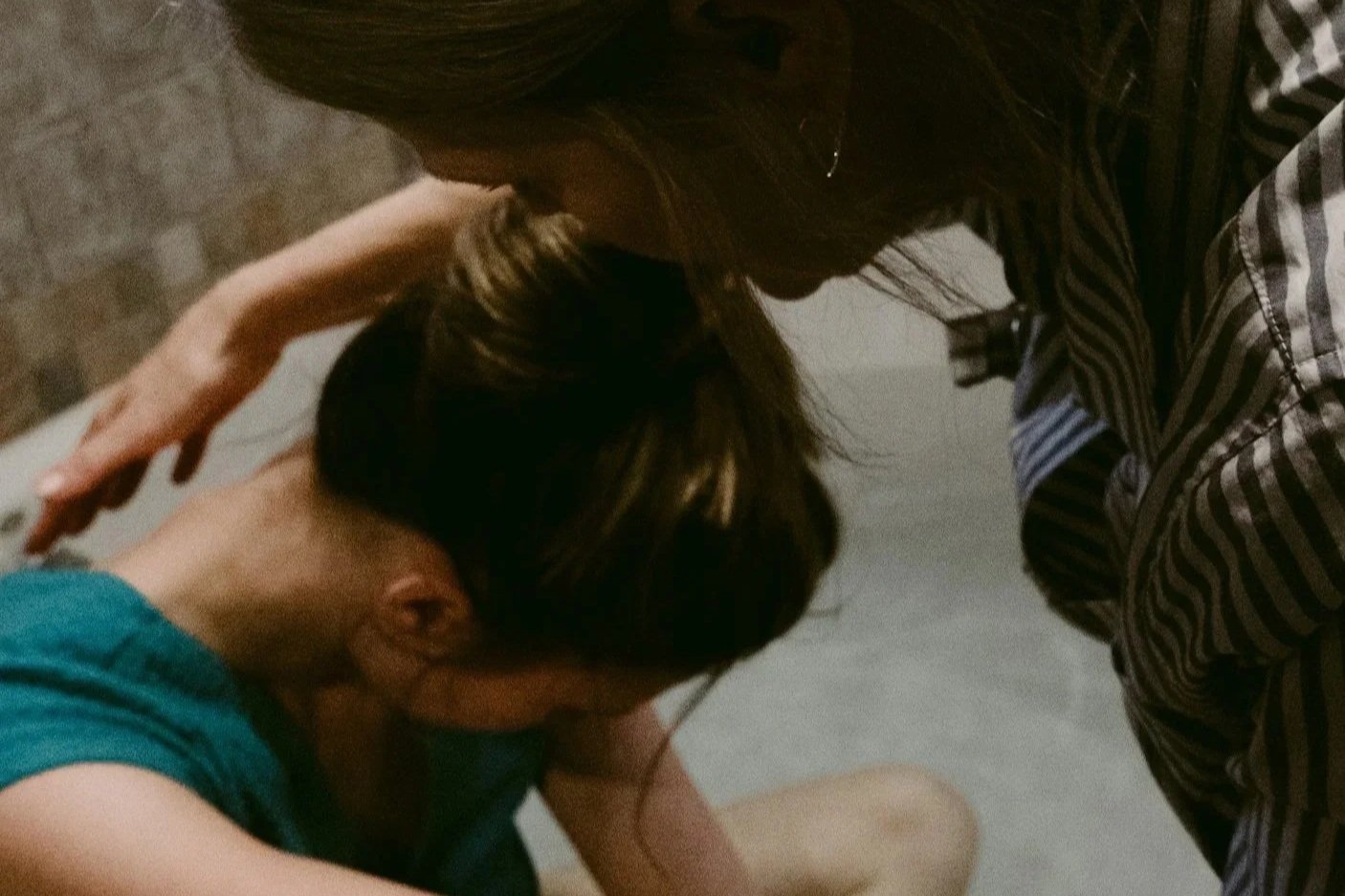 A woman helping a person in labor, comforting her, with their heads gently touching. The girl has short dark hair and is wearing a teal top, while the woman has long hair and is wearing a striped shirt.