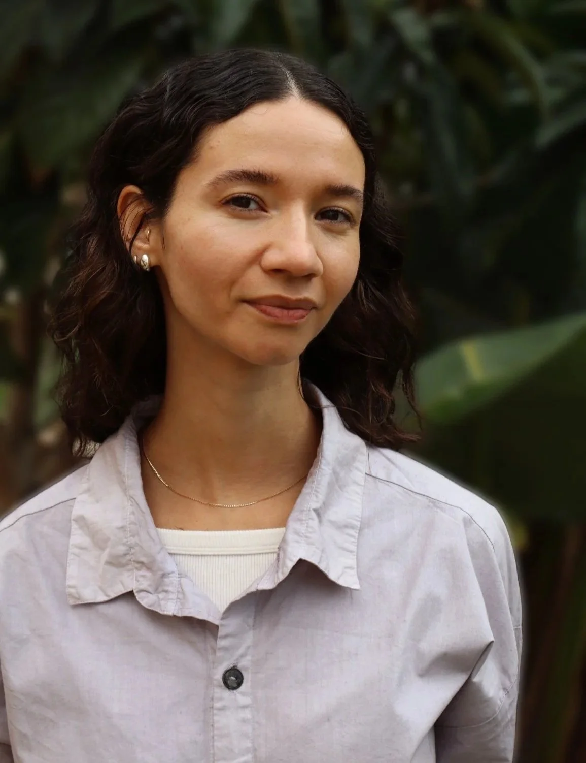 A young woman with curly dark hair wearing pearl earrings, a delicate necklace, a white shirt layered over a light-colored top, standing outdoors with green foliage in the background.