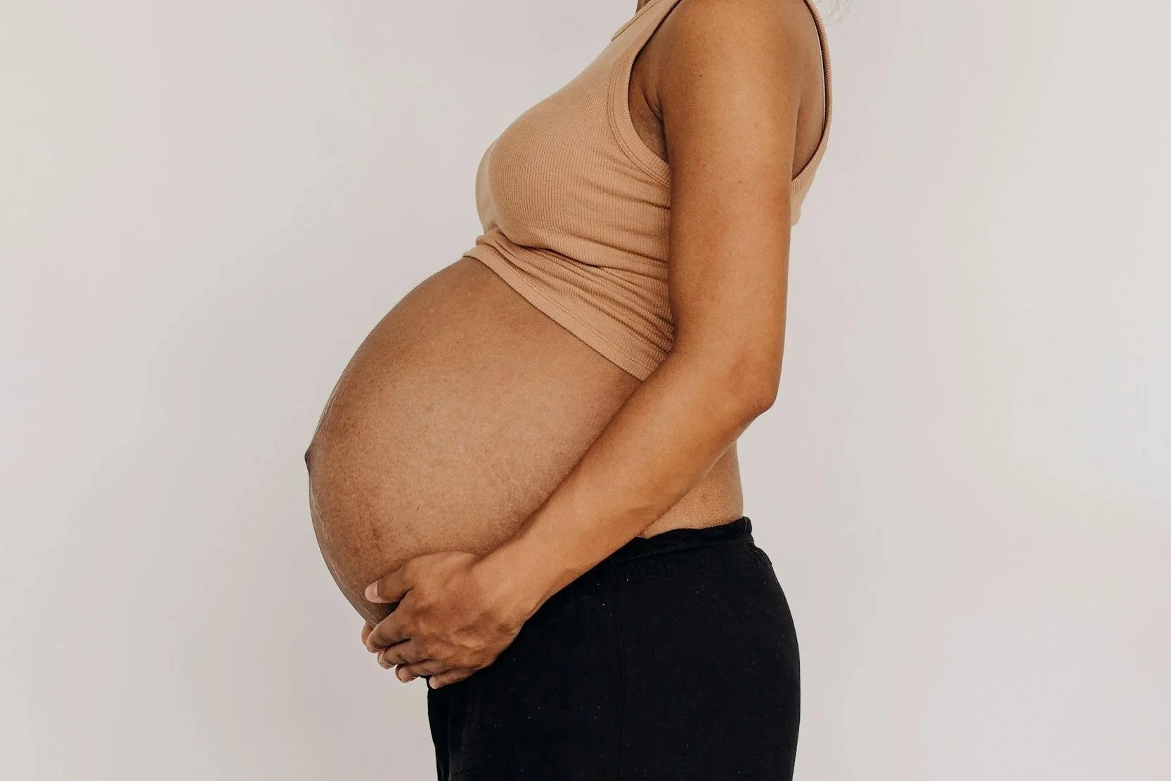 A pregnant woman wearing a beige tank top and black pants, holding her belly against a plain white wall.
