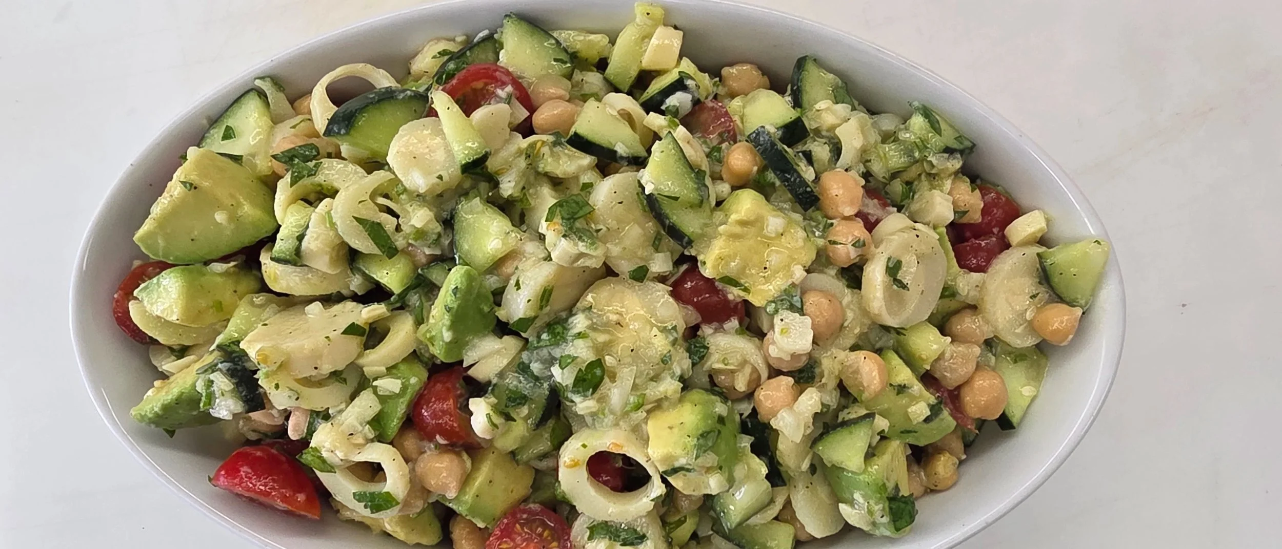 A bowl of hearts of palm salad featuring chopped cucumbers, cherry tomatoes, chickpeas, chopped herbs, and pasta, all served in a light dressing.
