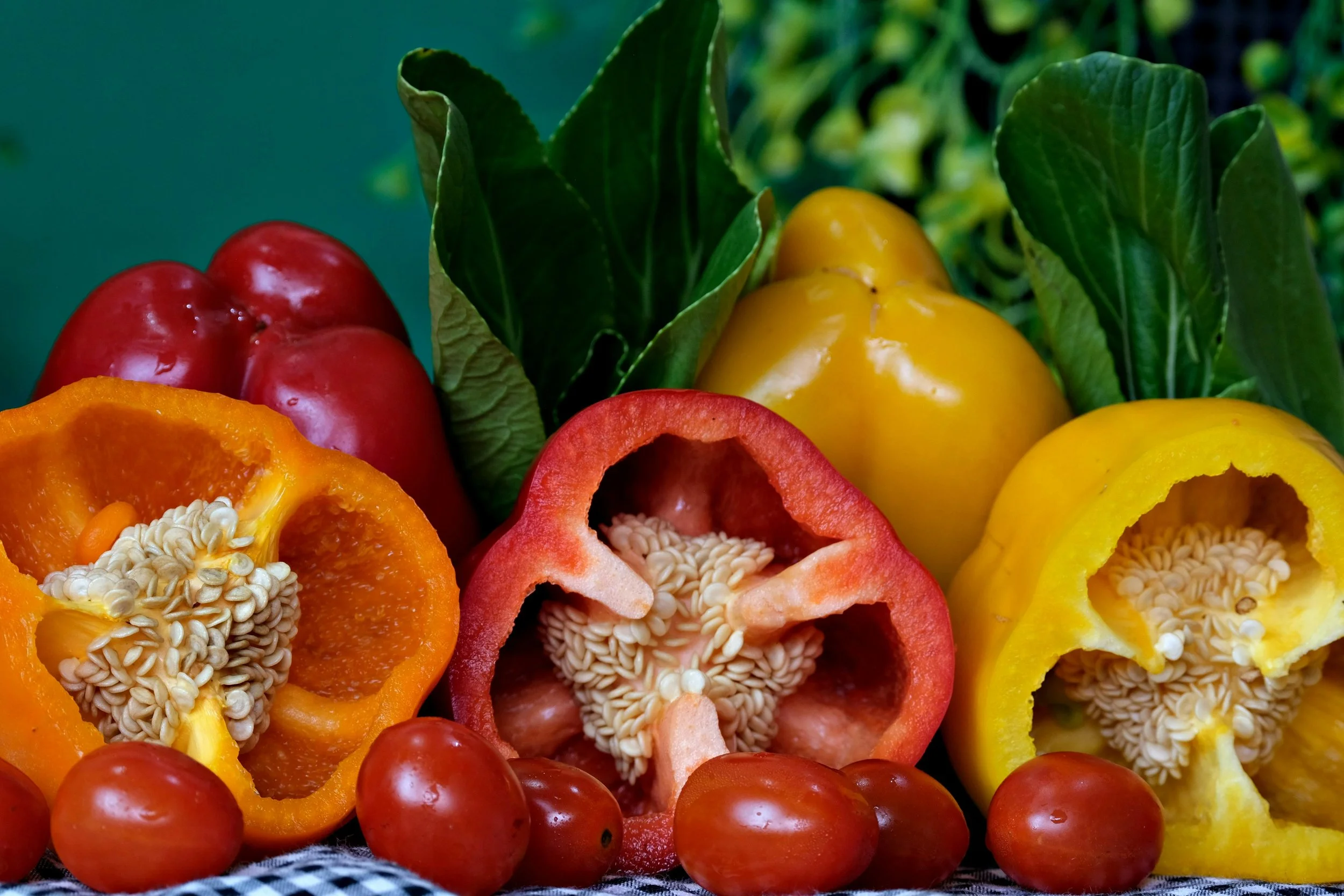 Various cut and whole bell peppers in red, yellow, and orange, with cherry tomatoes and green leaves in the background.