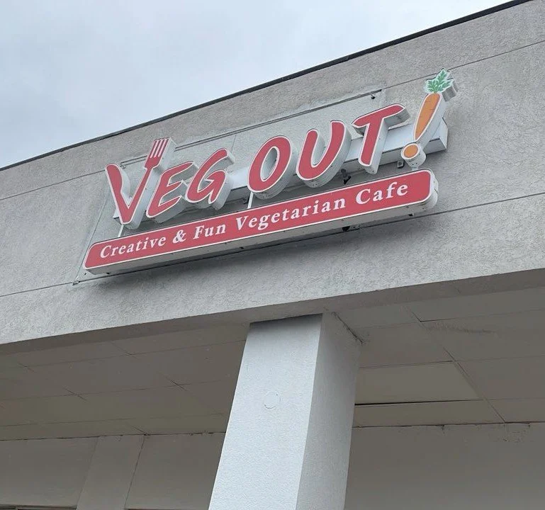 Sign for Veg Out Vegetarian Cafe with a fork and carrot logo, on a light-colored building exterior.