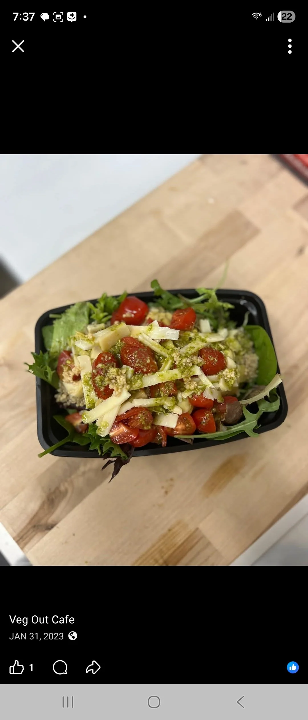 A fresh vegetable salad in a black container, consisting of cherry tomatoes, shredded cheese, leafy greens, and dressing.