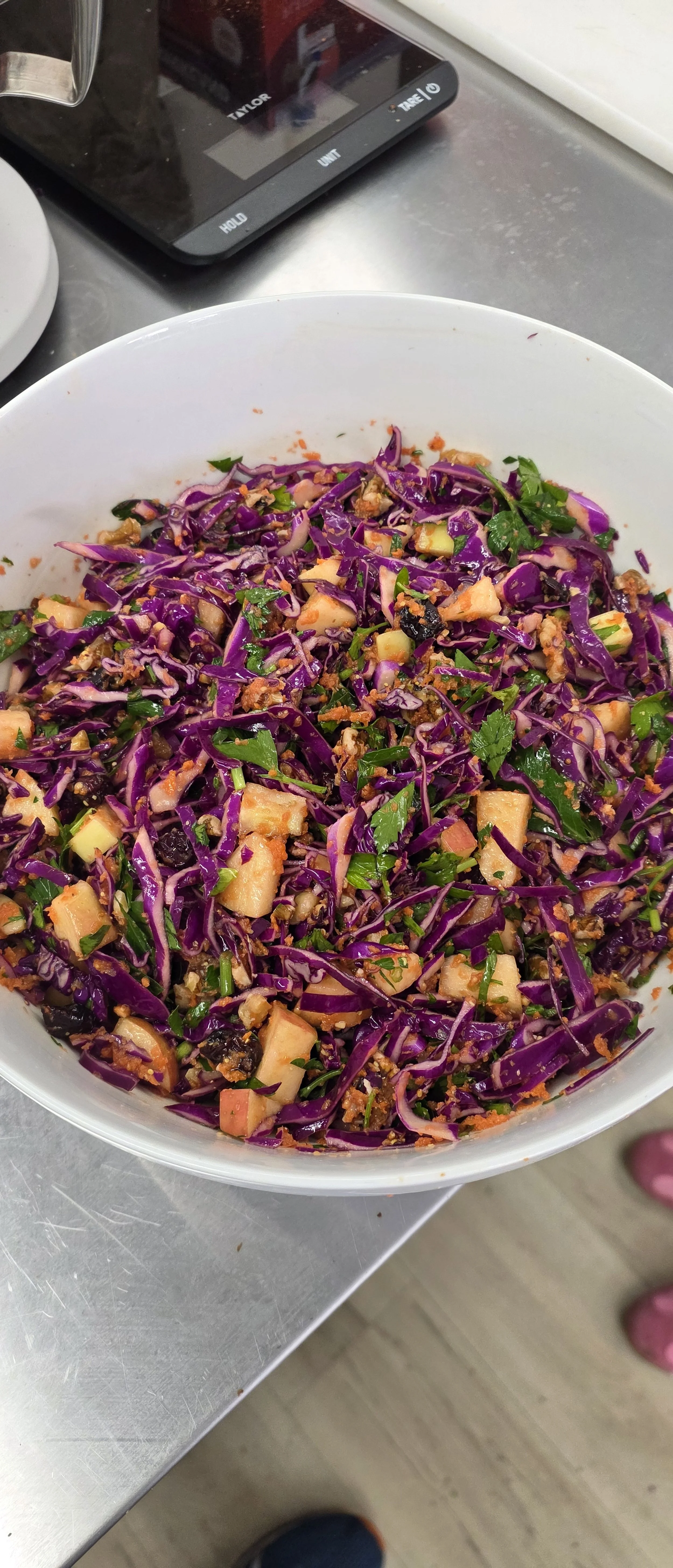 A colorful salad in a white bowl containing chopped purple cabbage, chopped green herbs, diced apples, and other vegetables, placed on a metal surface.