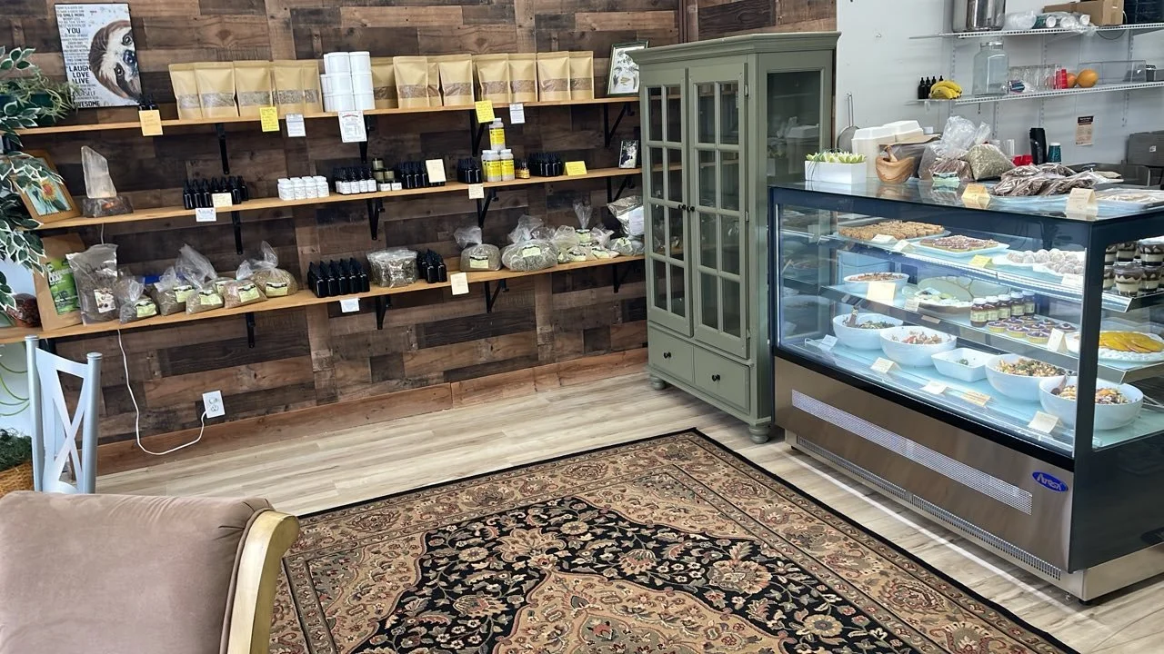 A cozy retail space featuring wooden shelves with various products like bottles and packaged goods, a glass display case filled with food items such as salads and desserts, a green wooden cabinet, a patterned area rug, and a partially visible beige a