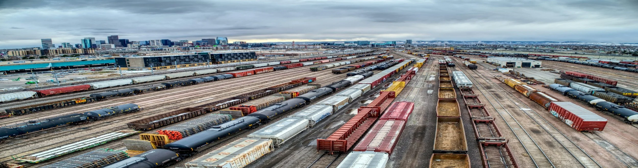 Overhead view of multiple train tracks with numerous freight cars and containers in a train yard, with a city skyline in the background under a cloudy sky.