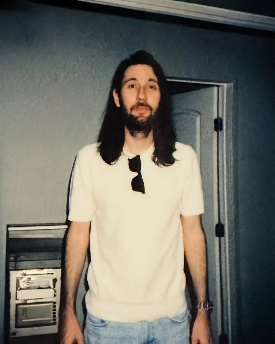 Man with long dark hair and beard, wearing a cream t-shirt with sunglasses hanging from the collar, standing in front of a closed door and a wall-mounted device.