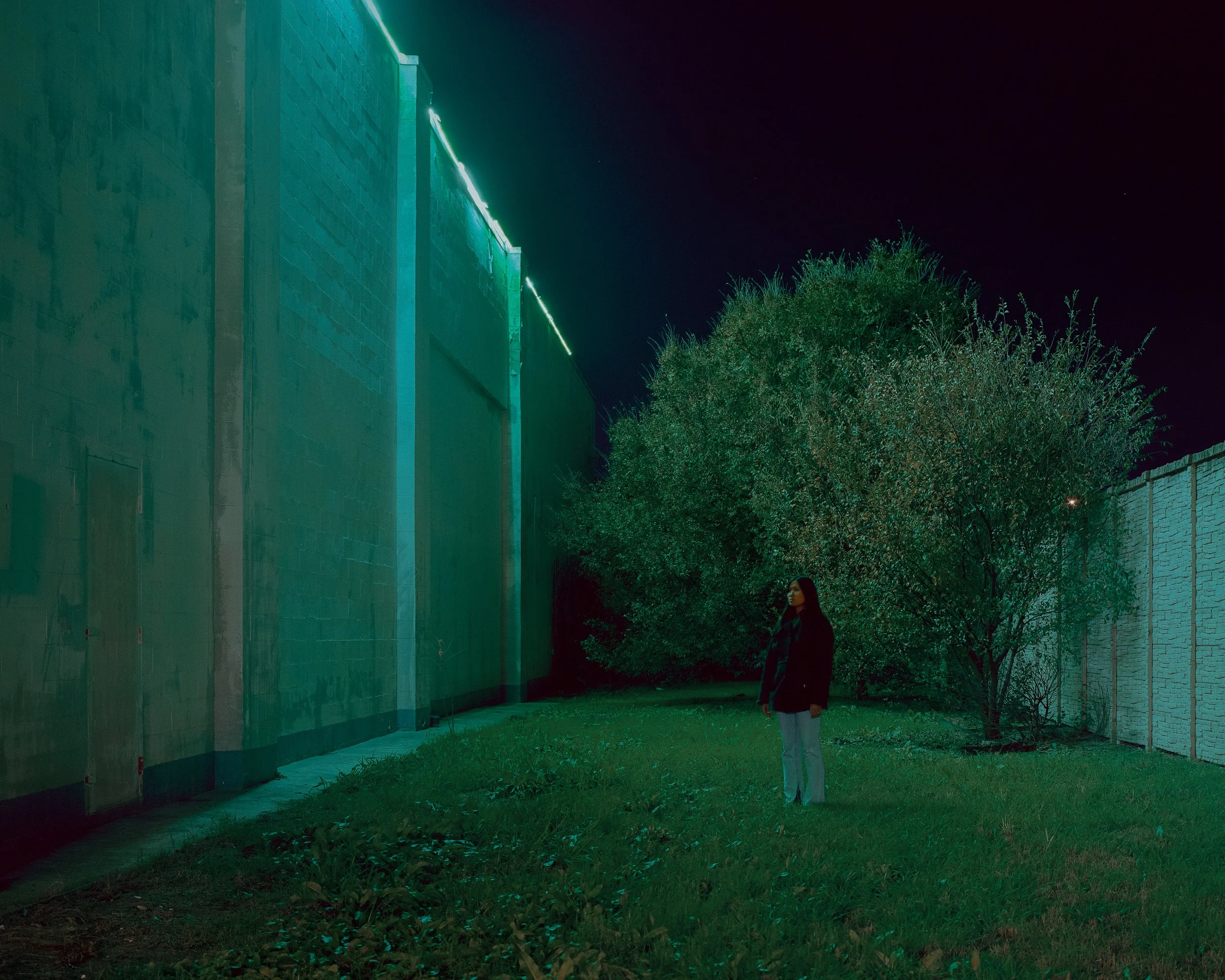 A woman standing on grass near a large tree and a brick wall, illuminated by green neon lights at night.