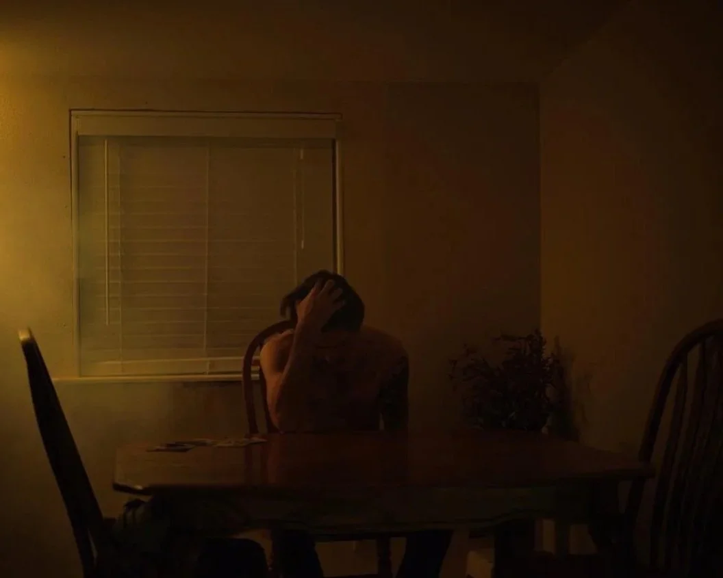 A person sitting at a dining table in a dimly lit room, with their head in their hand, appearing distressed or contemplative.