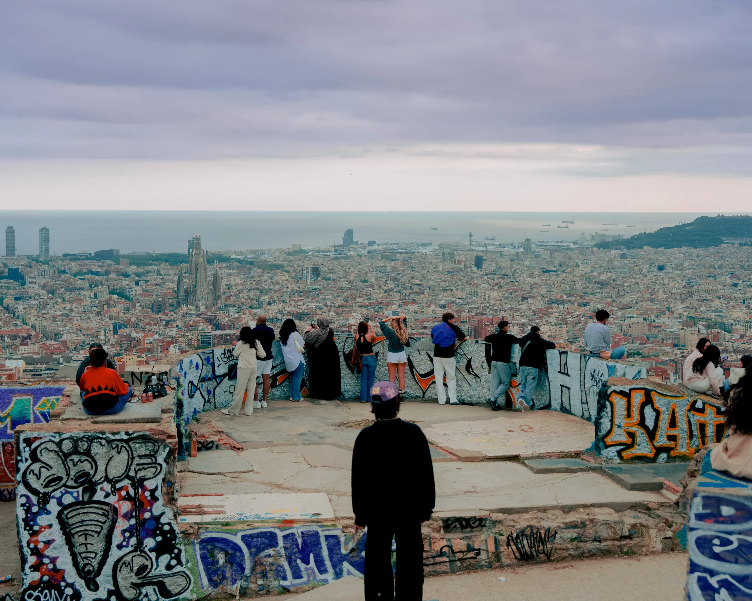 People sightseeing at the overlook of Barcelona, Spain with graffiti-covered walls, cityscape view, and the sea in the background, under cloudy sky.