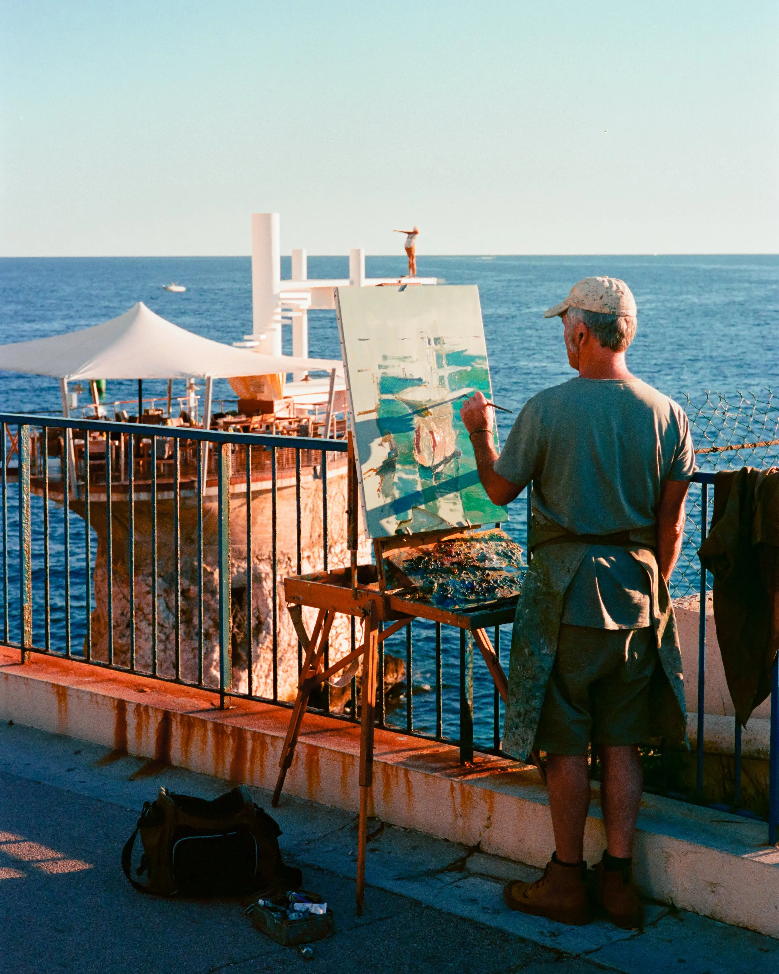 A male artist painting on a canvas outdoors by the ocean, with a boat and a person standing on a platform visible in the background.