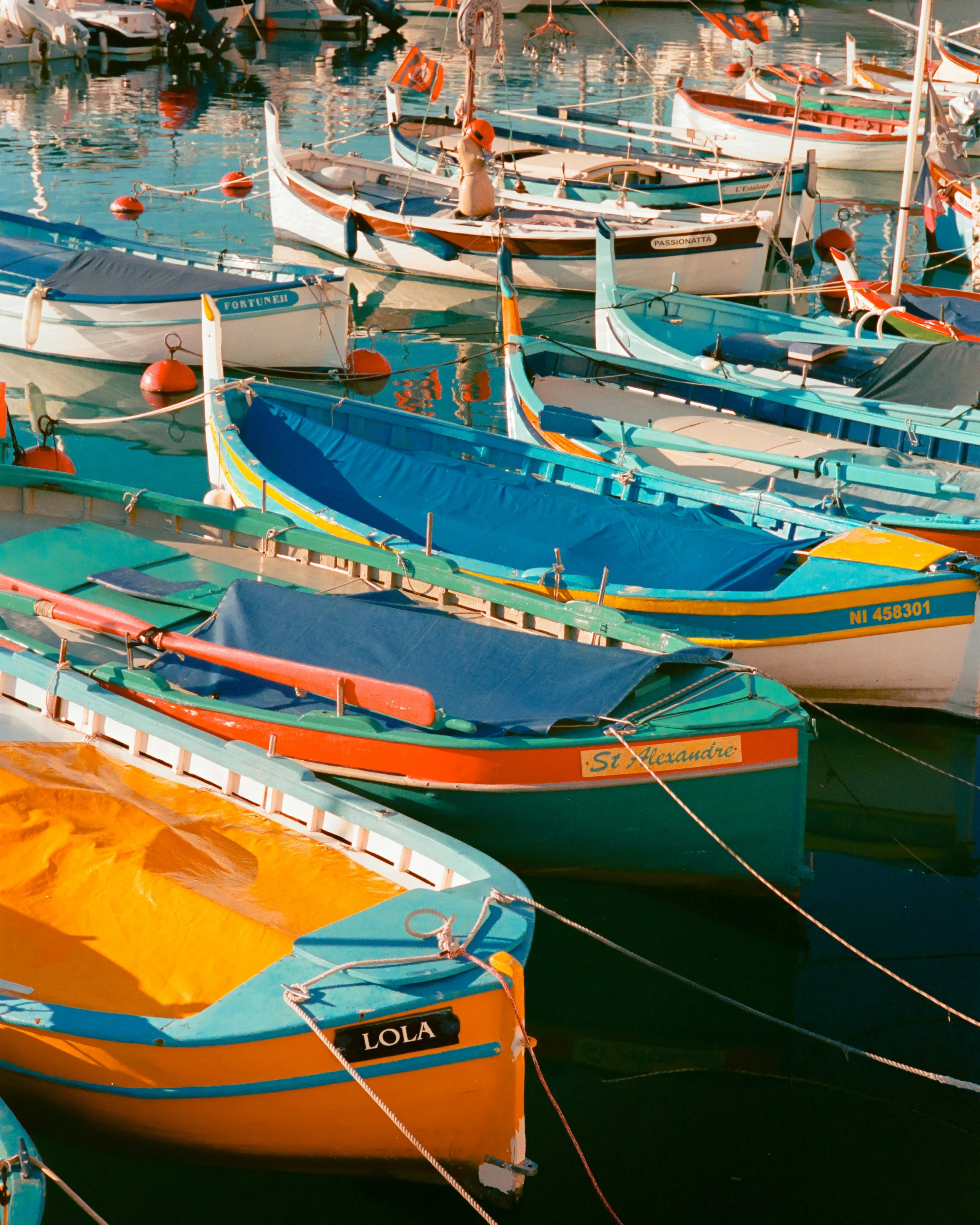 Colorful boats are docked at a marina on calm water. The boats are in various colors including blue, orange, and white, with some covered and others uncovered. They are moored with ropes and orange buoys.