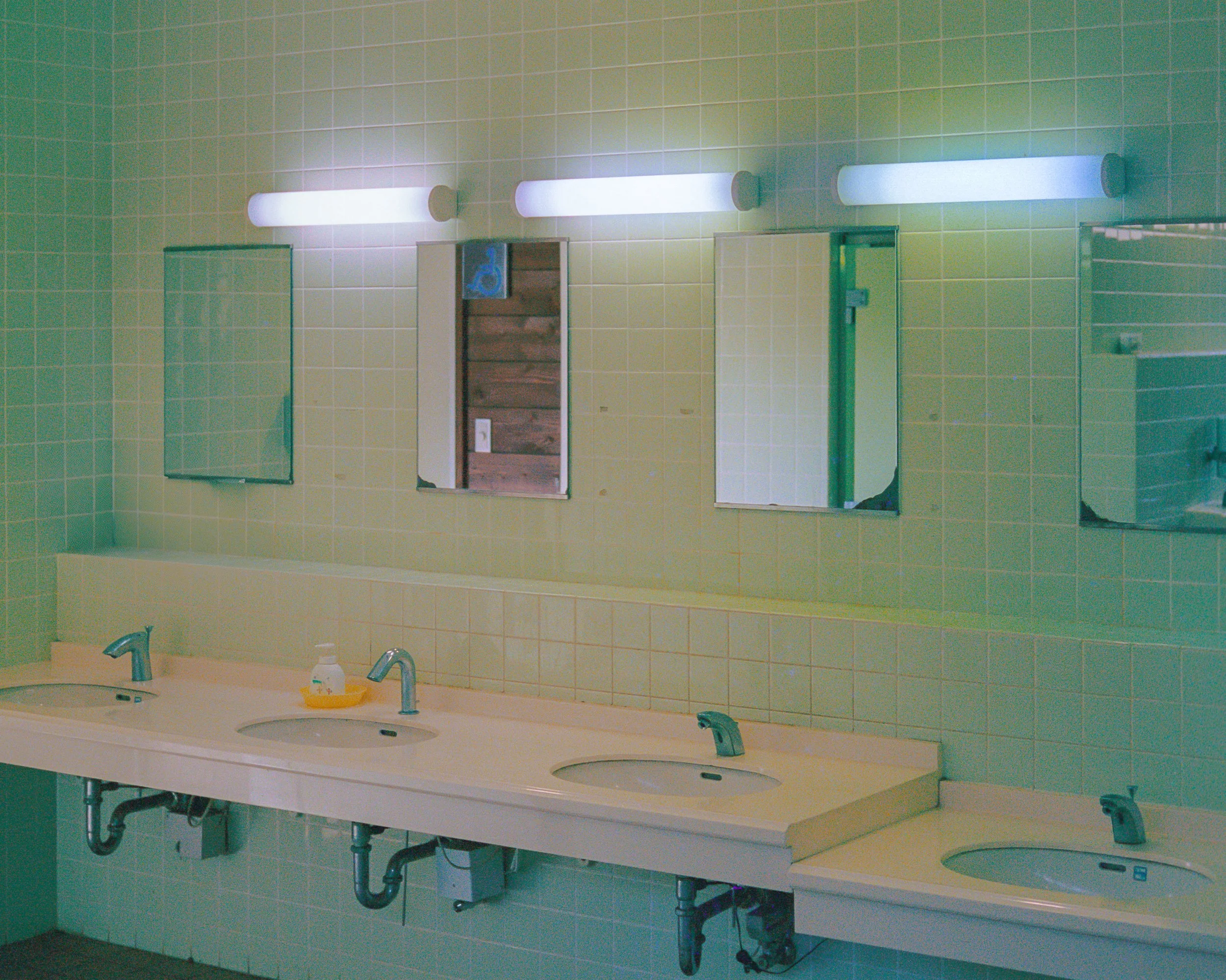 Public men's bathroom with four sinks, three mirrors, and a soap dispenser on the left.
White countertop, green-tiled wall, with toilet paper dispenser visible in the mirror on the left.
Lighting fixtures above the mirrors, with a door visible in the