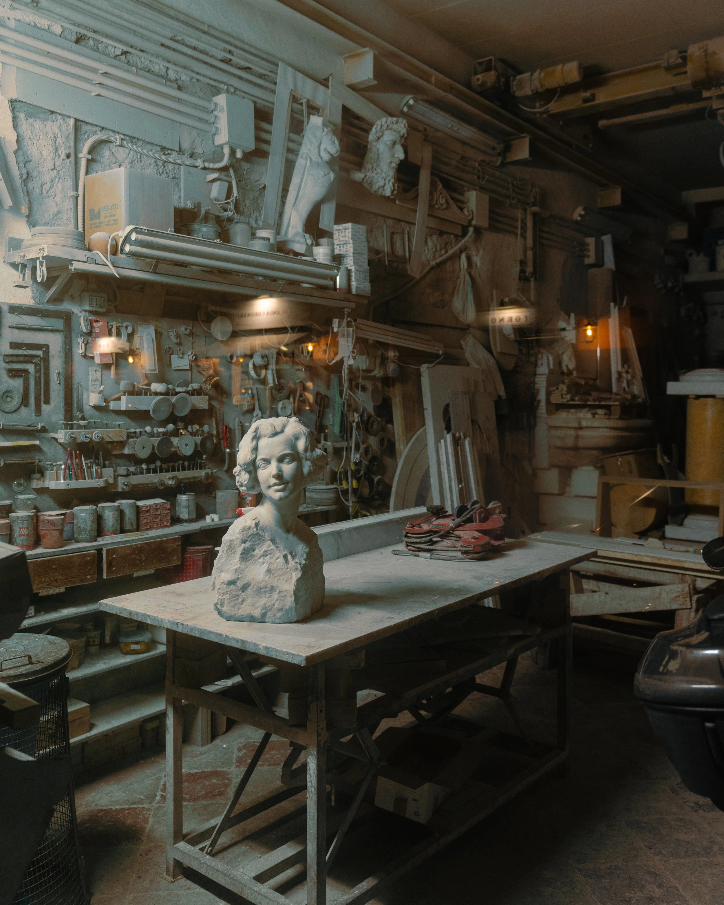 A sculpture workshop with a bust of a woman on a wooden table, surrounded by various tools and sculptures, including busts of male figures on shelves and walls.
