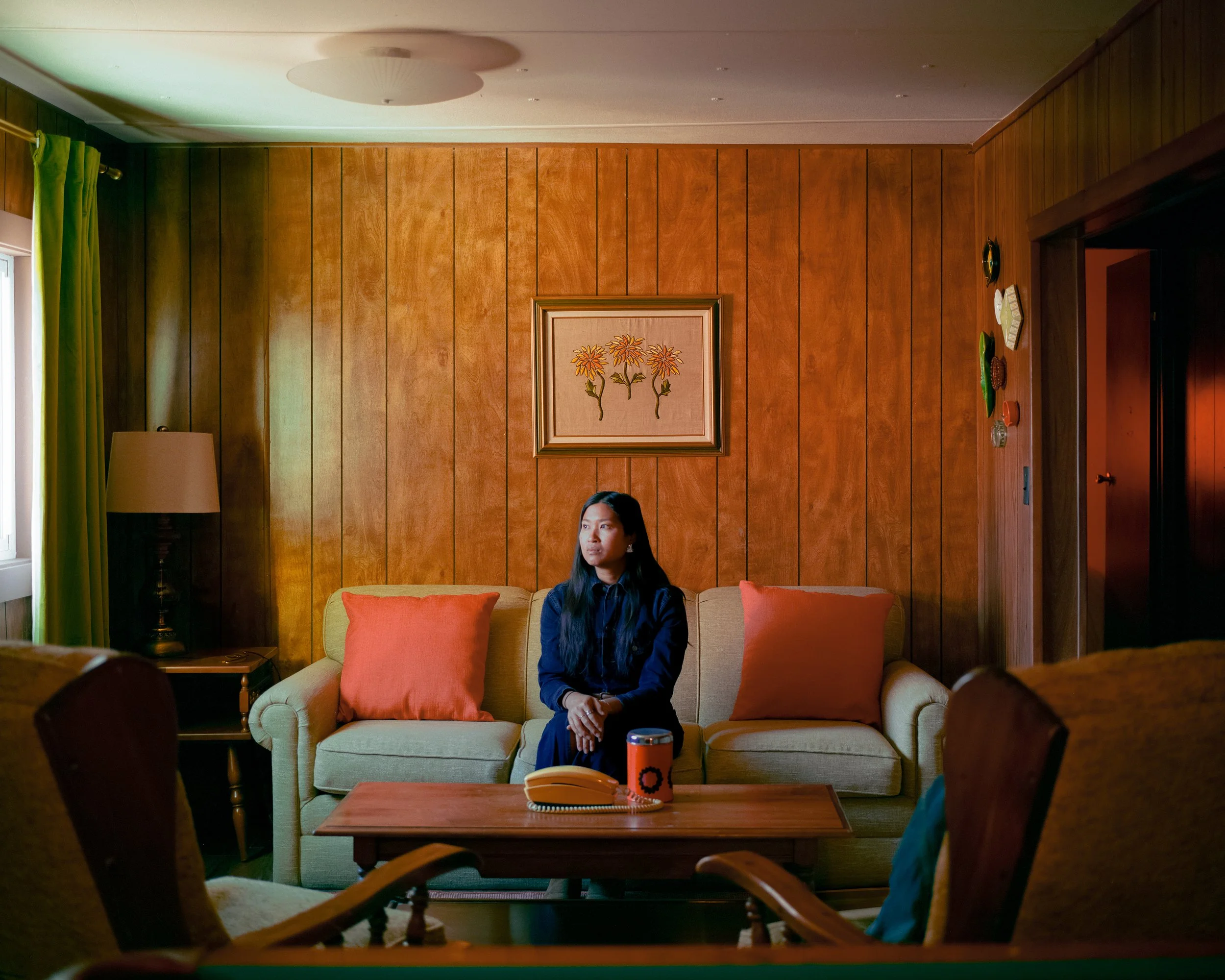 A woman with long black hair sitting on a beige sofa with orange pillows in a wood-paneled living room, with a picture of flowers hanging on the wall behind her, a side table with a lamp and phone, a wooden coffee table with a yellow phone and a blac