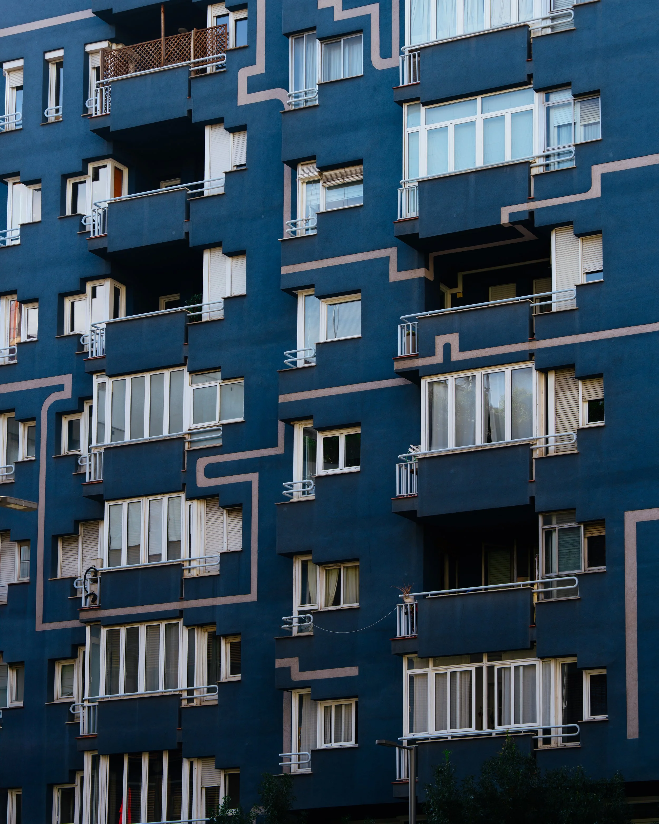 Close-up of a blue apartment building with multiple windows and small balconies, some with plants and various window coverings.