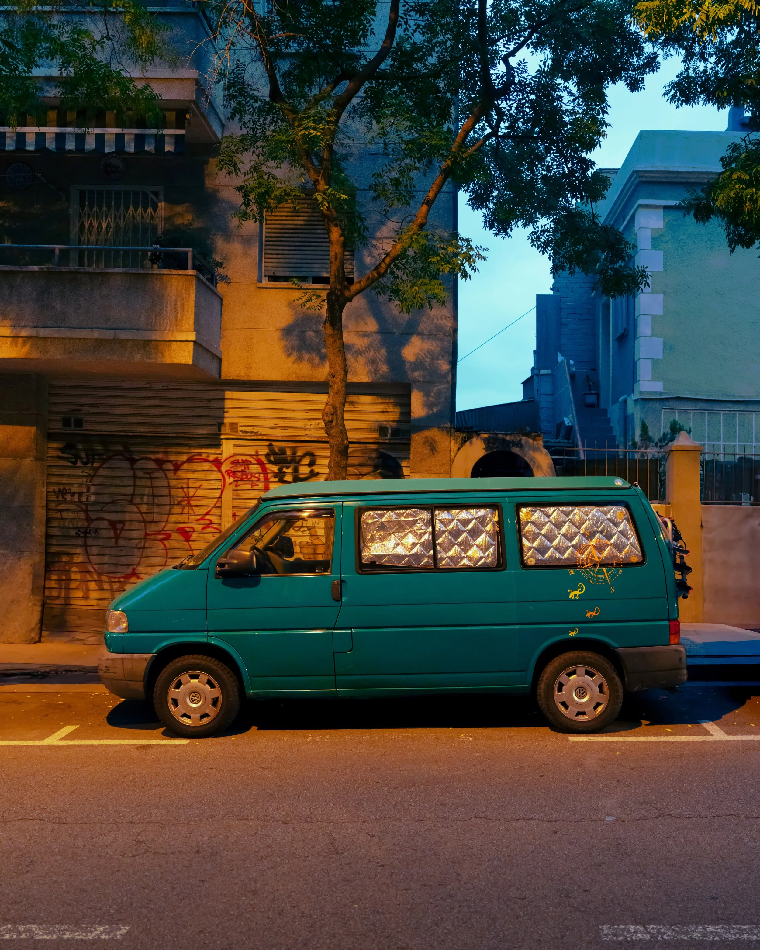 A teal-colored van parked on a city street at dusk with a tree behind it. The van has reflective diamond-patterned covers over the windows and graffiti on a metal shutter nearby.