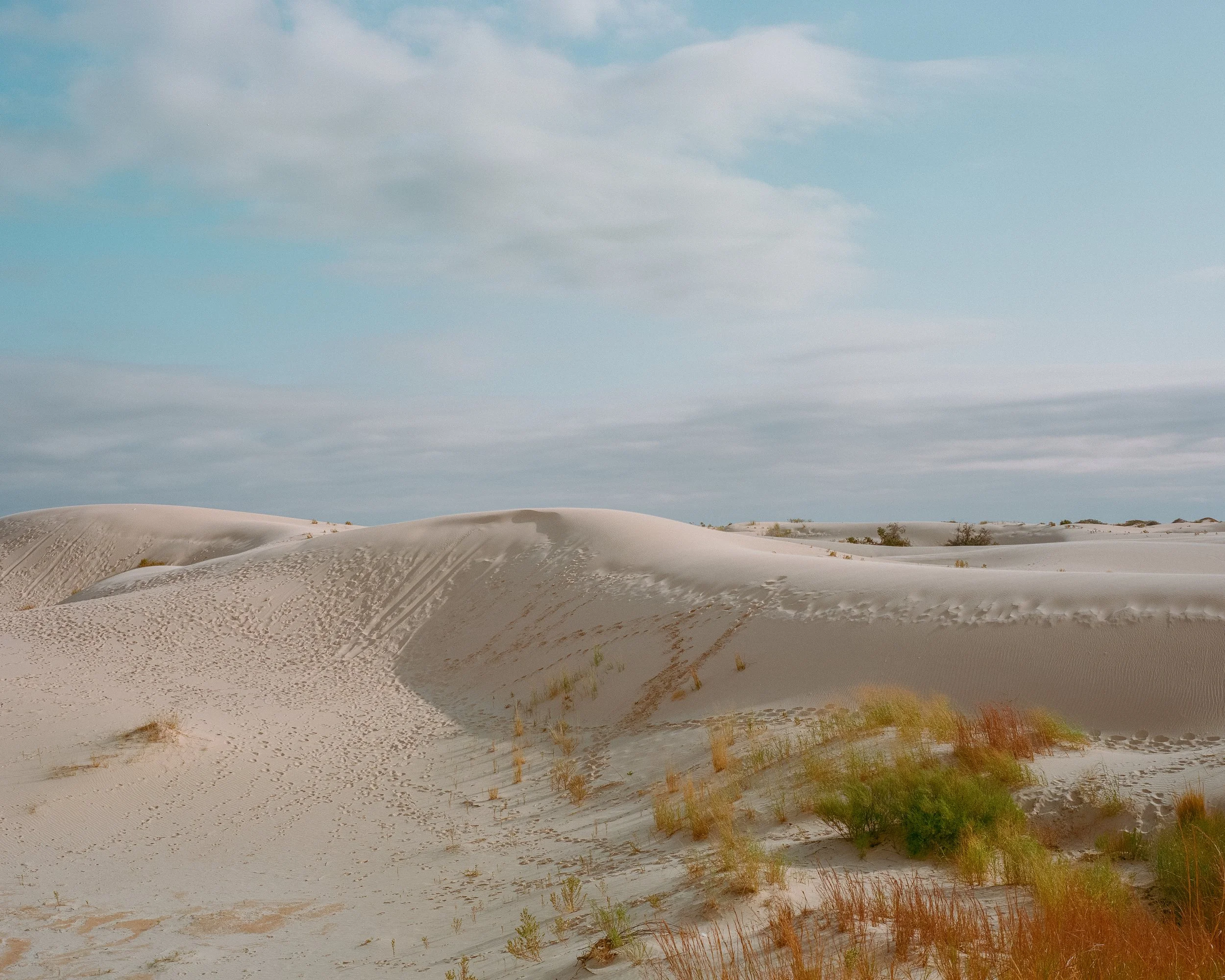 Landscape of sand dunes with sparse vegetation under a partly cloudy sky.
