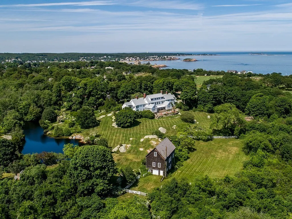 Aerial view of a large white house surrounded by greenery, a pond, and open lawns with another smaller dark house nearby, overlooking a coastal town and ocean in the background.