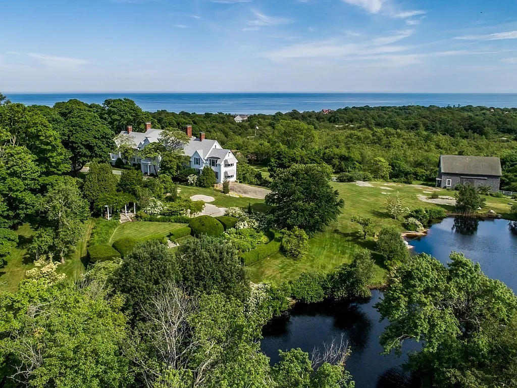 A scenic view of a coastal landscape with a large white house, lush green trees, a pond, and a blue ocean in the background.