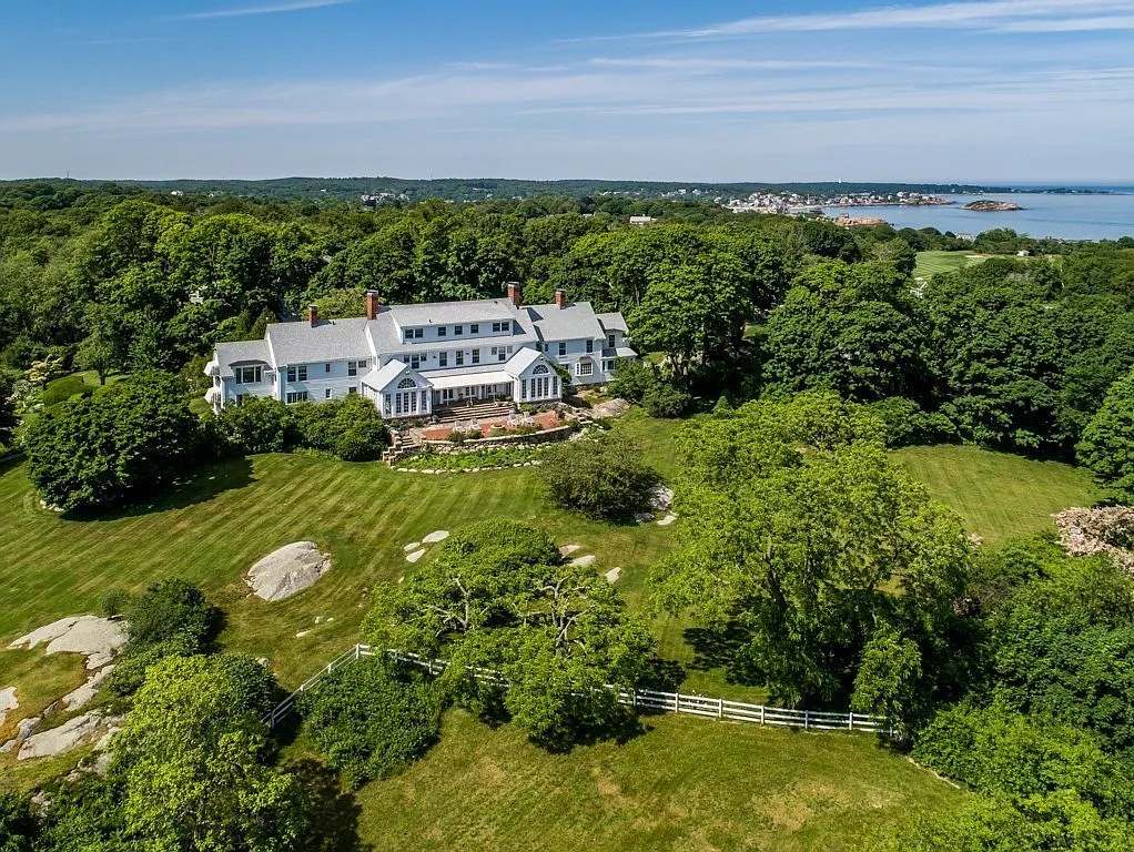 Large white house with multiple levels and chimneys, surrounded by lush green trees and a well-manicured lawn, overlooking a body of water in the distance.