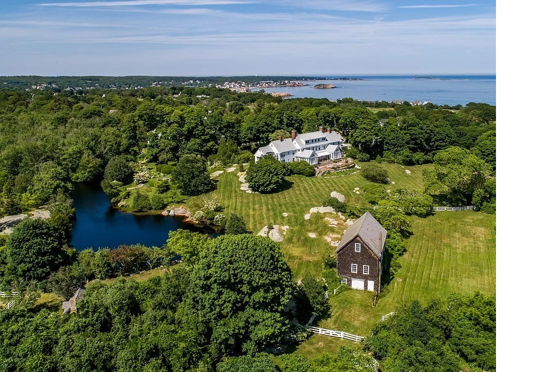 Aerial view of a large white house with a gray roof surrounded by trees, a pond, and a grassy lawn, with the ocean in the background.
