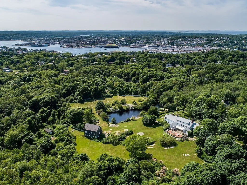 Aerial view of a green landscape with trees, houses, a pond, and a large white house, overlooking a body of water with a city skyline in the background.