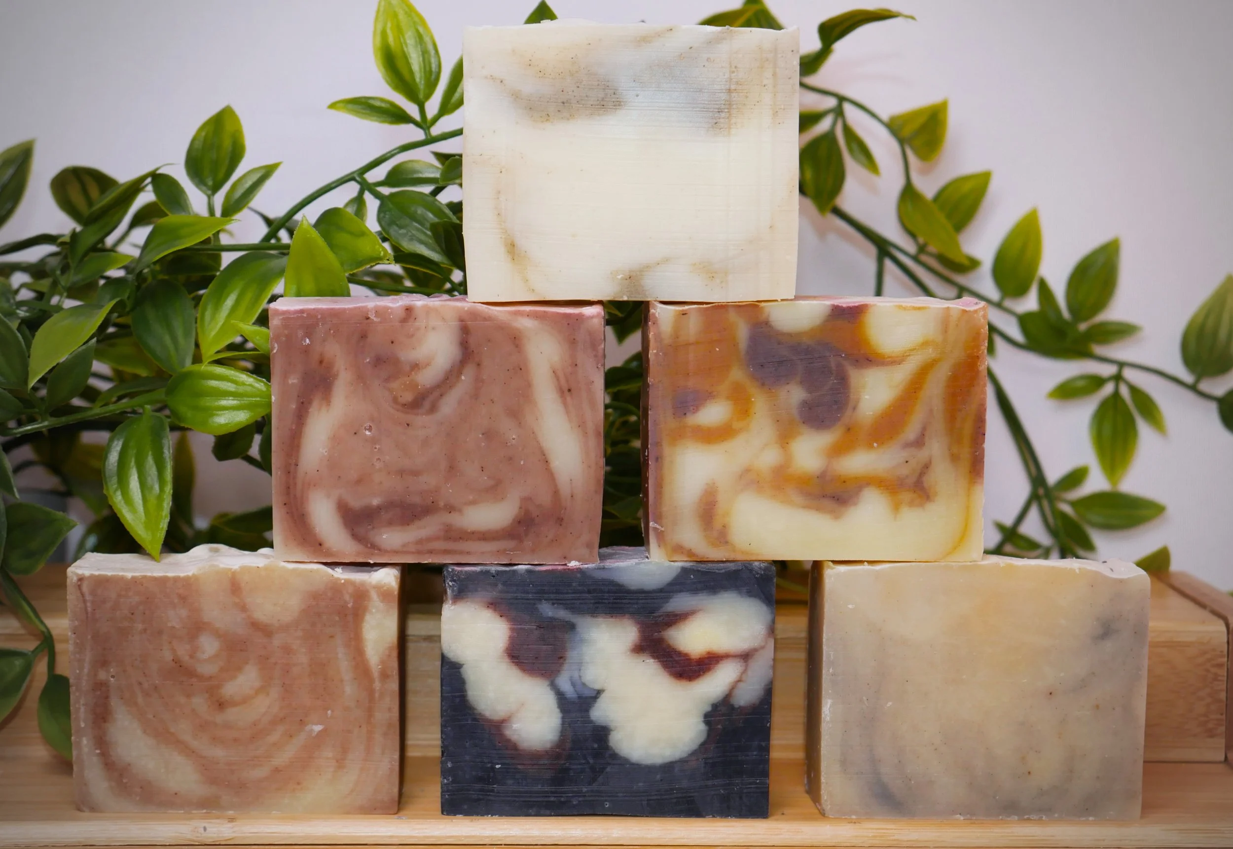 A collection of seven handmade soap bars with various beautiful  marbled patterns, arranged on a wooden tray with green leafy plants in the background.