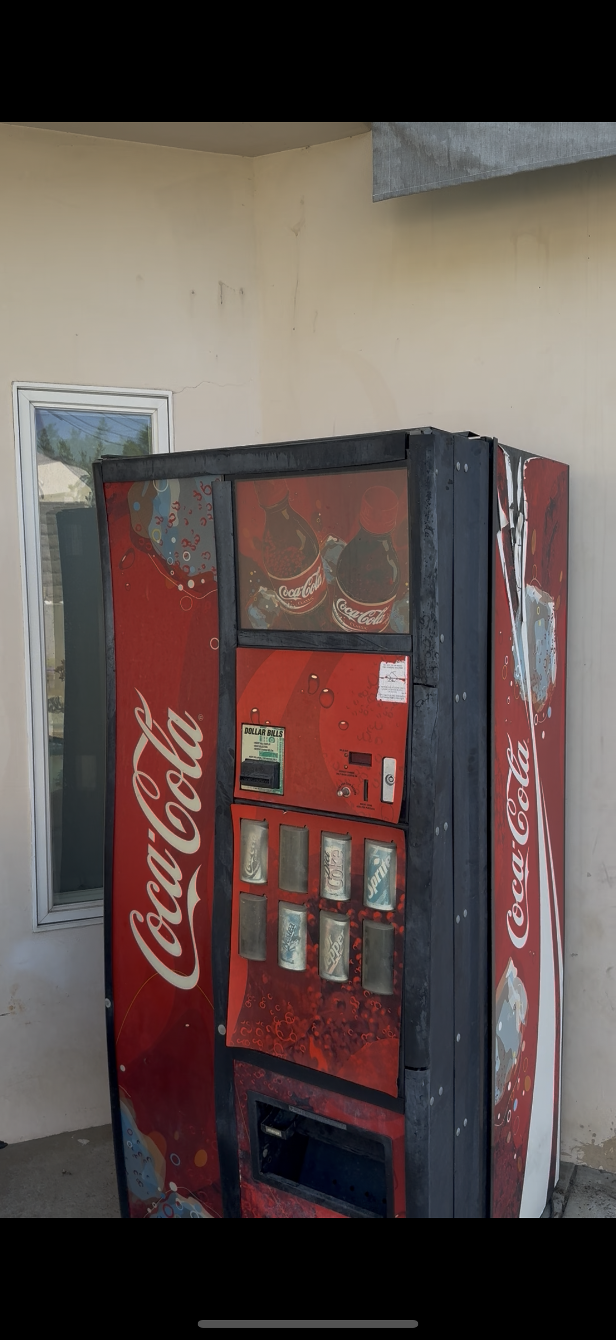 A Coca-Cola vending machine outside a building with a window, against a beige wall.