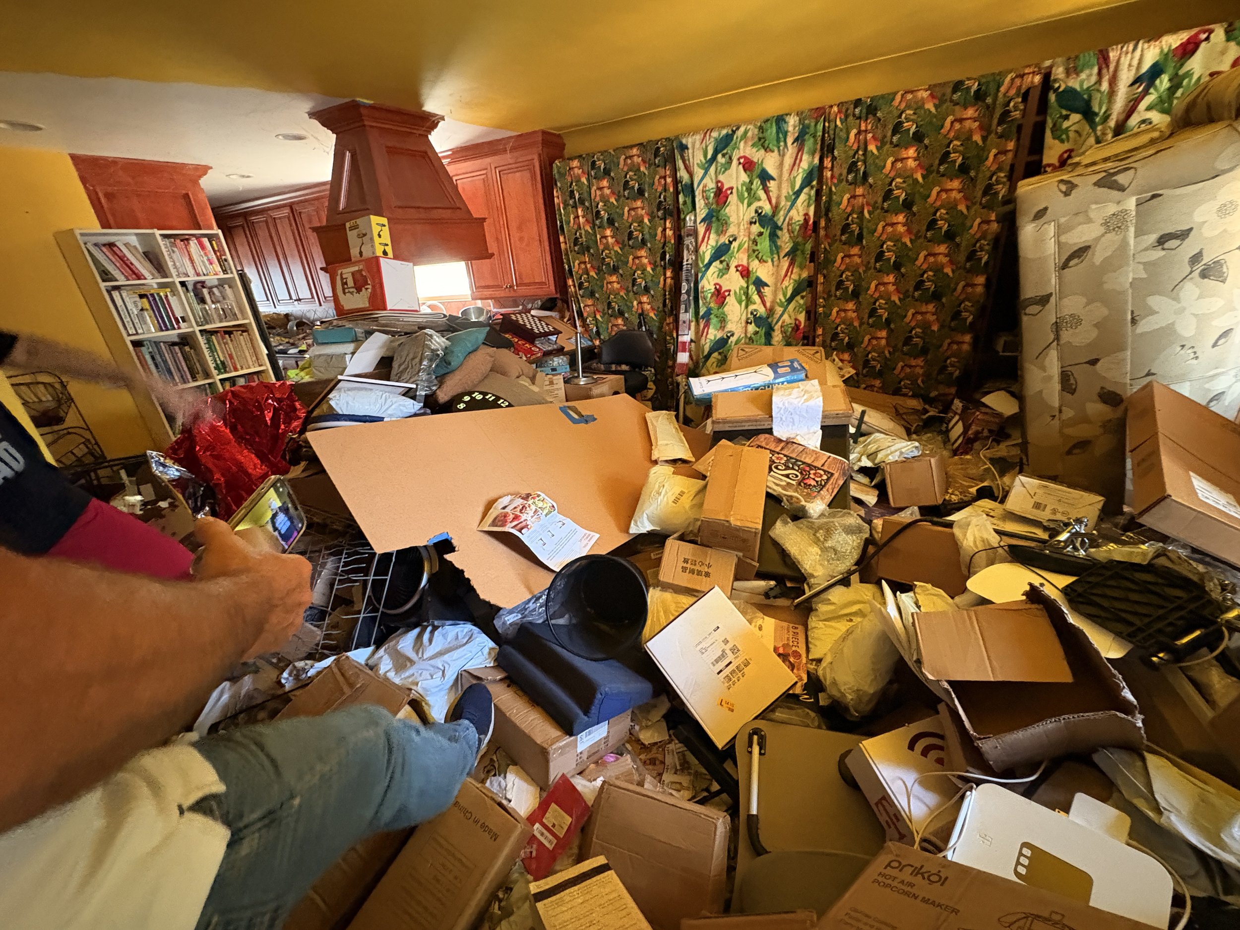 A cluttered room with boxes, bags, and various household items scattered across the floor, with bookshelves and curtains in the background.