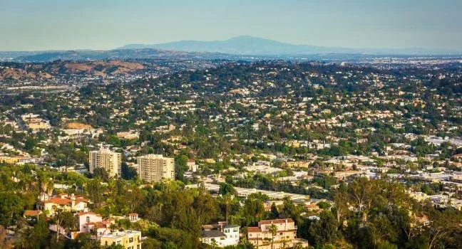 Cityscape view of a large urban area with many buildings, trees, and mountains in the distance under a clear sky.