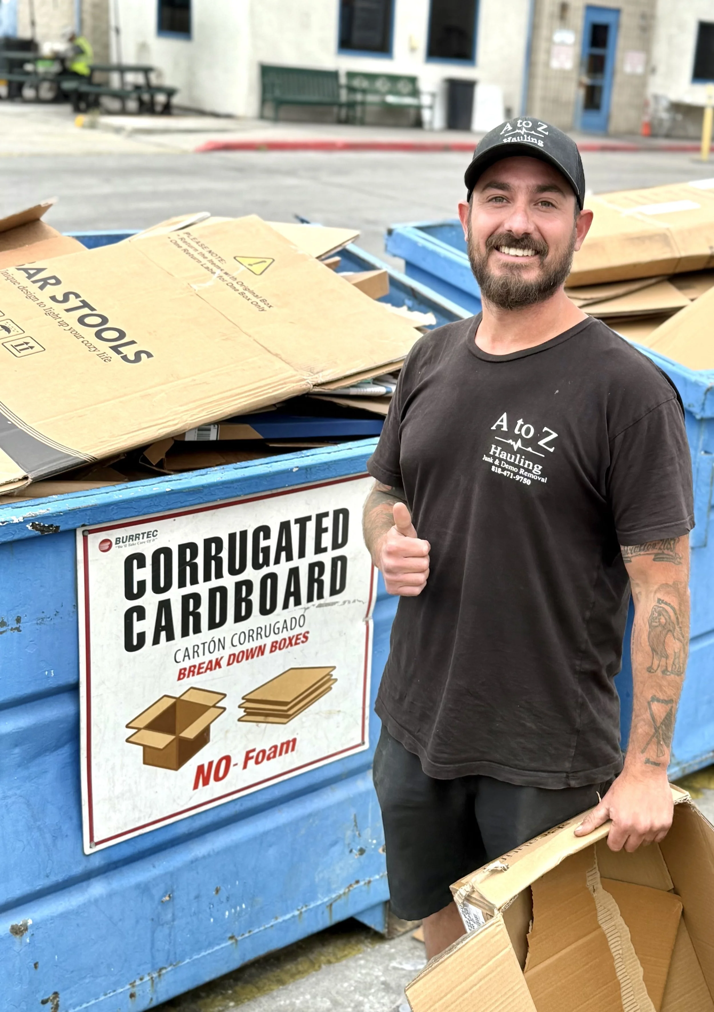 holding a piece of cardboard in front of a blue dumpster. He is wearing a black T-shirt and a black cap with white text. Behind him, there is a sign on the dumpster reading 'CORRUGATED CARDBOARD' and describing it as non-foam and breakable for recycling.