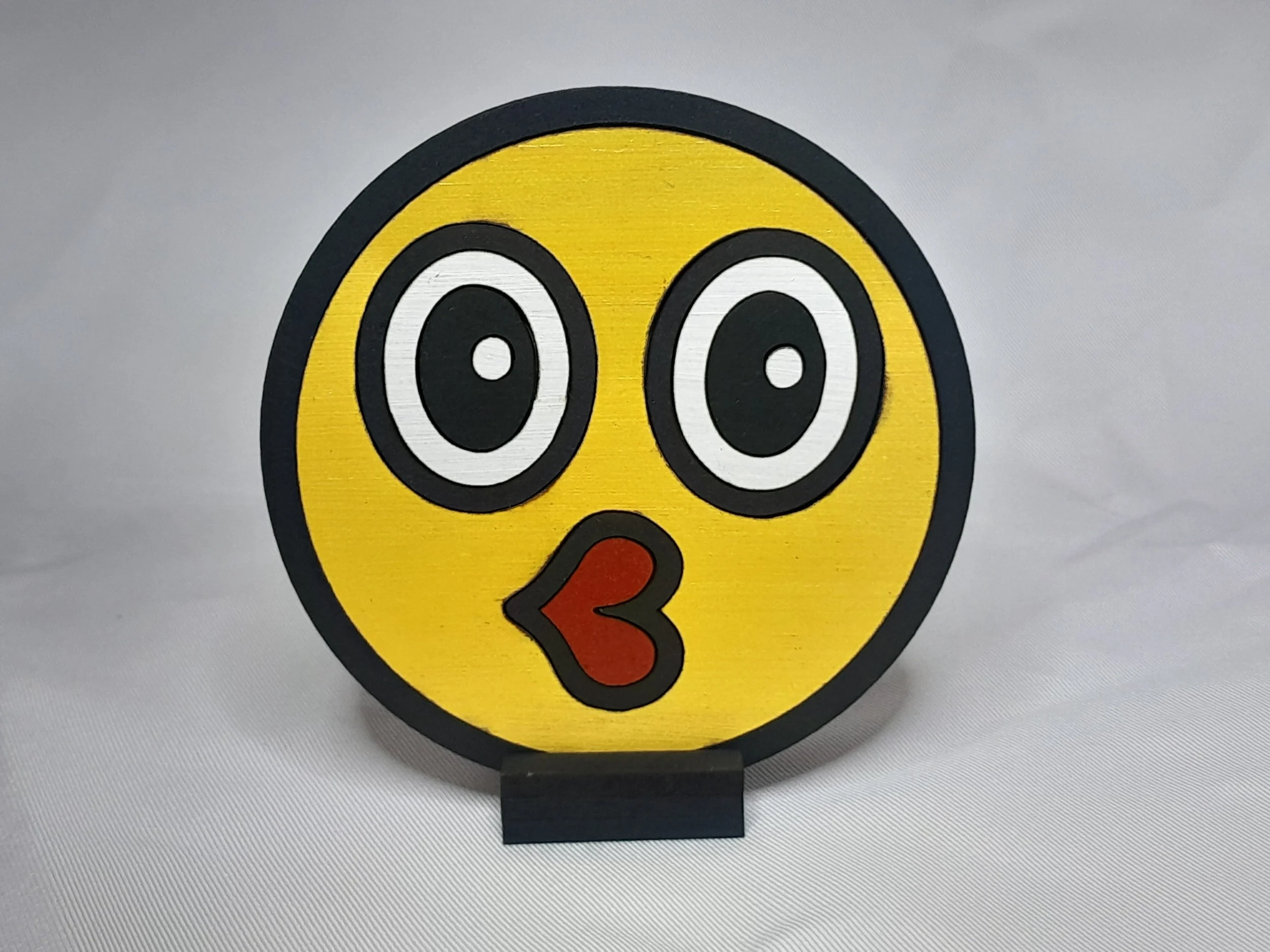 “Wide‑eyed kissy face emoji magnet with bold cartoon eyes and red puckered lips, laser‑cut painted wood on a small display easel.”