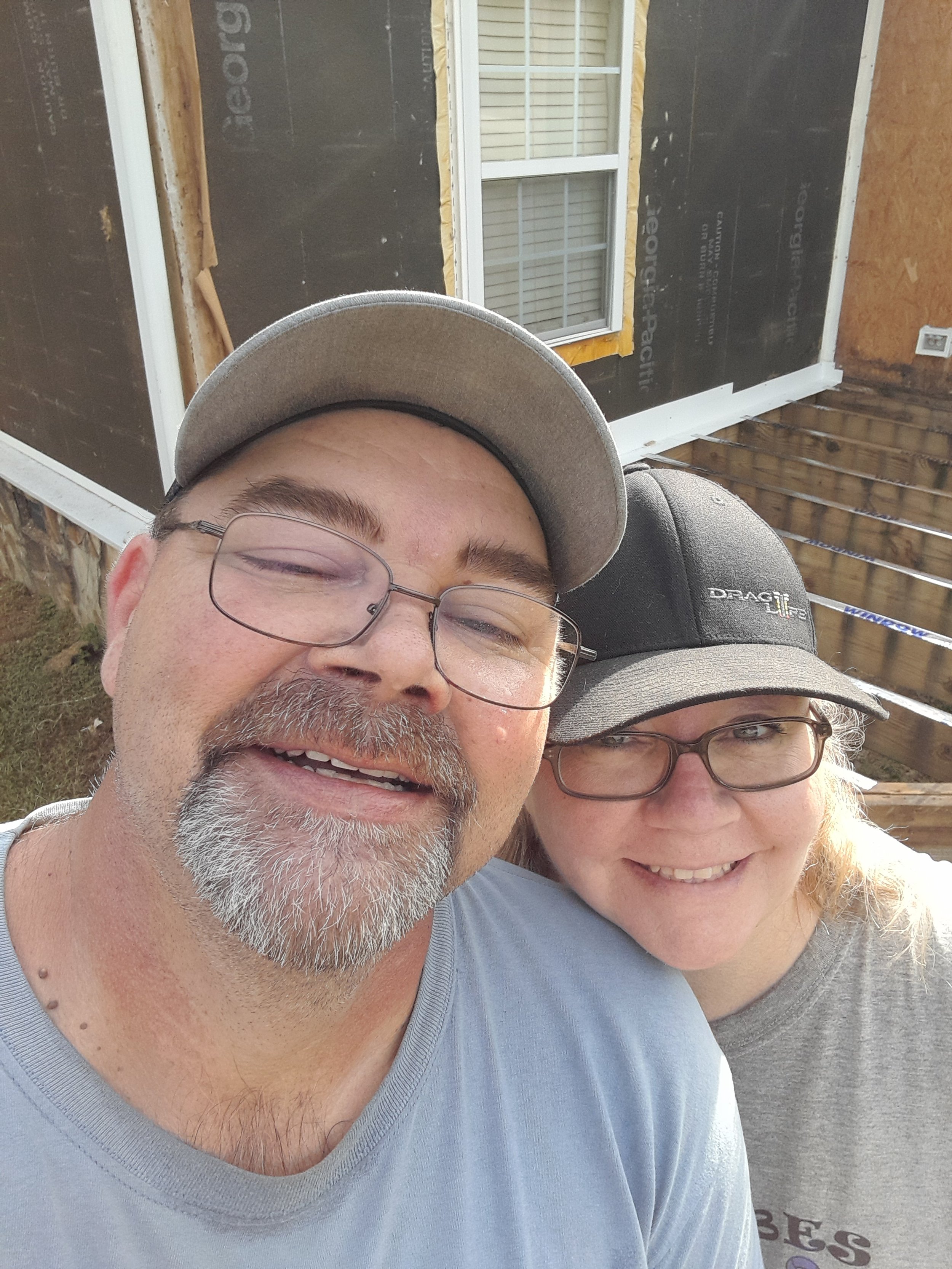 Close-up photo of a man and woman smiling outdoors, wearing glasses and caps, with a house under construction in the background.
